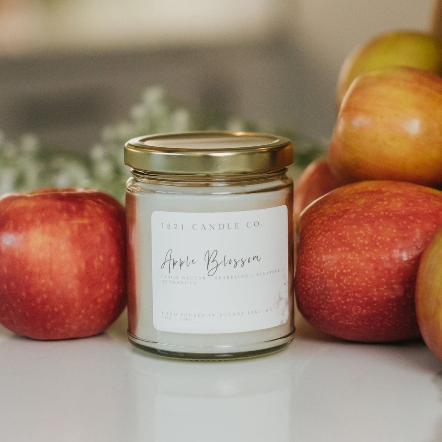 Apple Blossom soy blend candle next to fresh apples and baby's breath flowers on a white countertop.