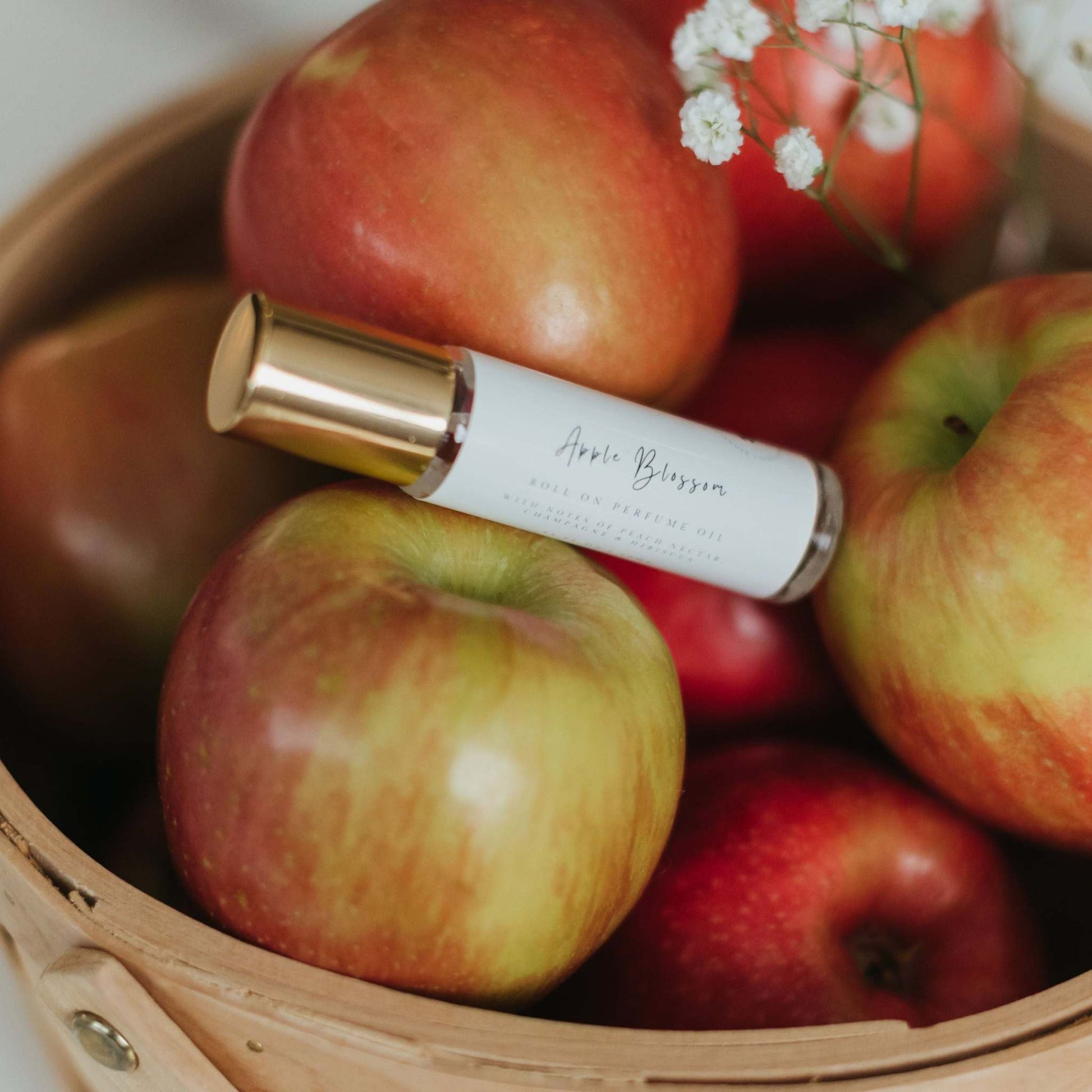 A glass bottle of Apple Blossom roller ball perfume oil with gold cap rests on a basket filled with fresh red apple, accompanied by delicate white baby's breath flowers. The scene evokes a crisp and floral fragrance, perfect for a fun and refreshing everyday scent.
