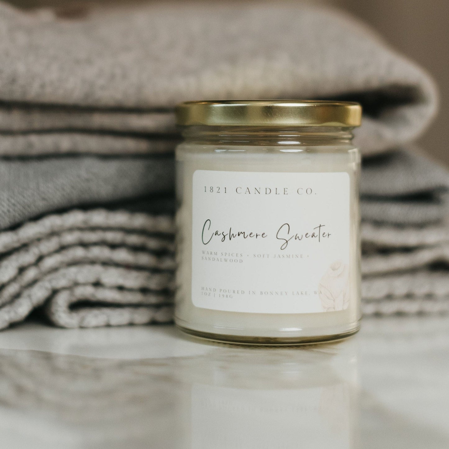 Phthalate free soy blend candle called Cashmere Sweater sitting on a neutral toned sweaters. A cozy gift for slow living and soft comforting scents.