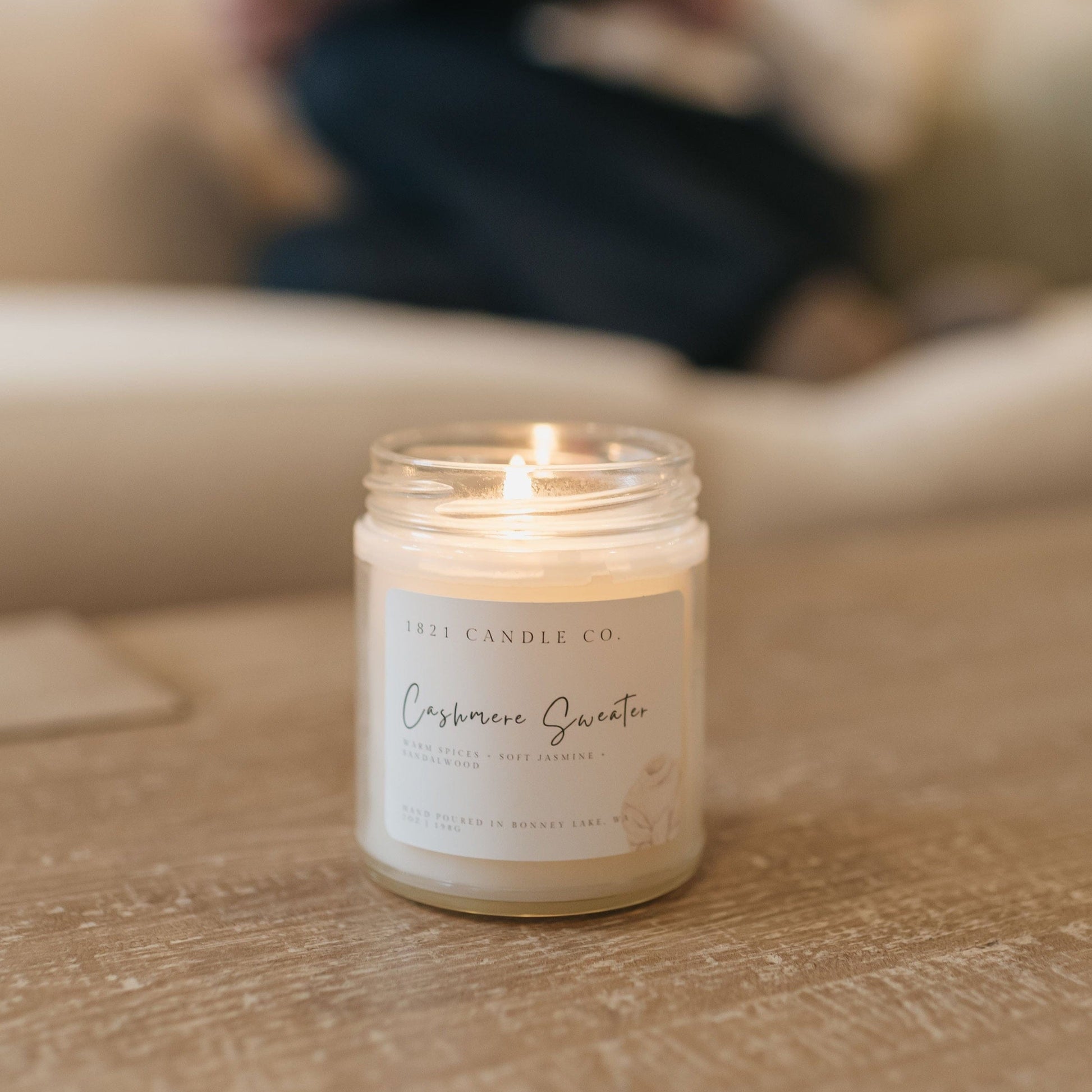 Cashmere Sweater Soy Blend Candle lit that is sitting on a coffee table with a person reading in the background in a sofa.