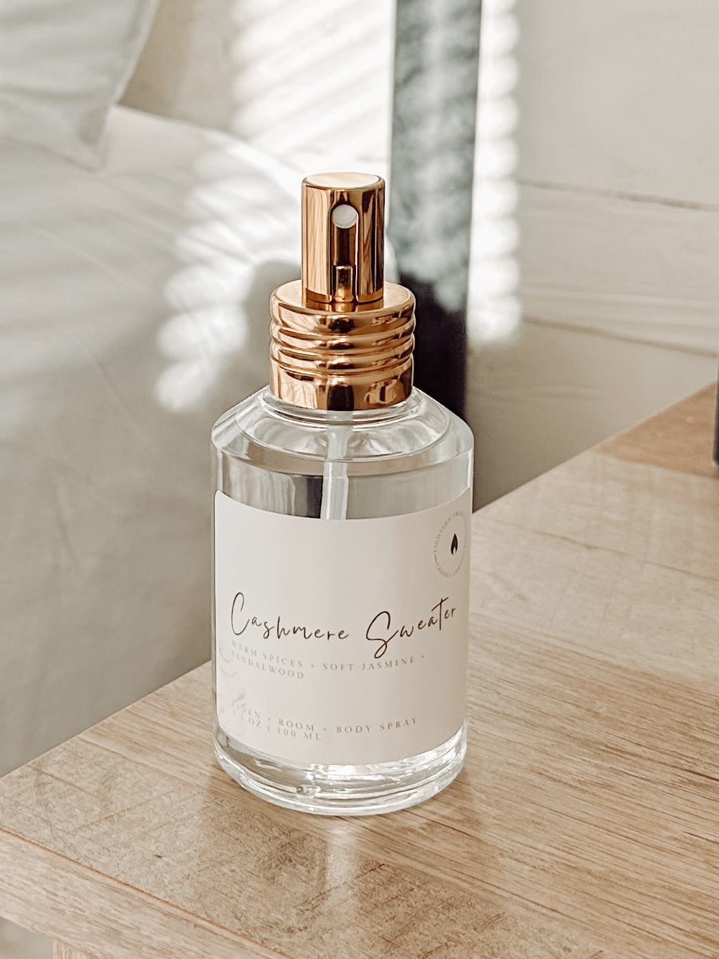 Cashmere Sweater linen + room spray by 1821 Candle Co in clear glass bottle with gold lid sitting on a bedside table with a bed in the background.