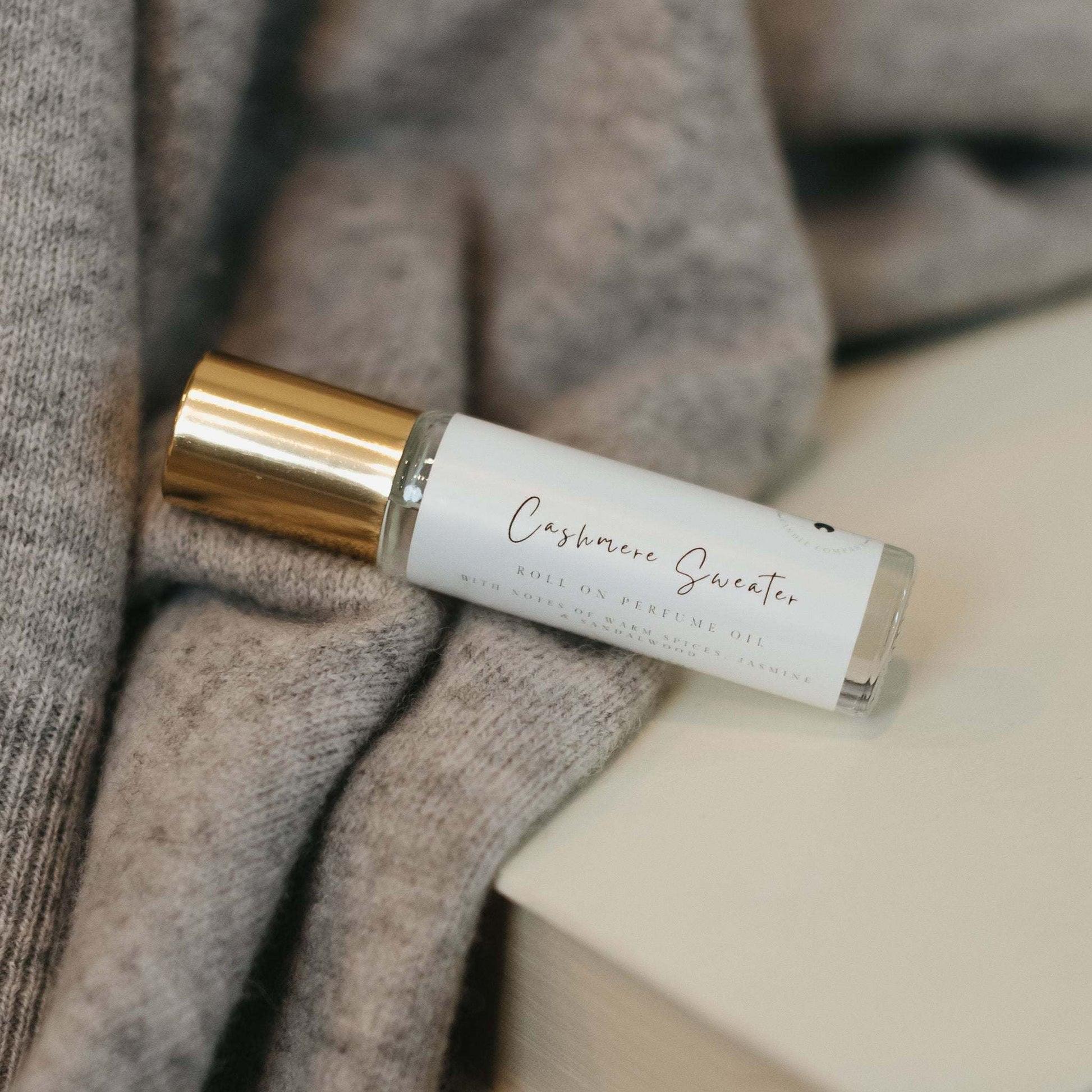 A Cashmere Sweater roller ball perfume rests on a soft gray sweater, placed atop a stack of neutral-toned books. The minimal, cozy aesthetic enhances the feeling of warmth and comfort, perfectly reflecting the fragrances soft notes of cedarwood, musk and jasmine, This elegant and travel friendly perfume oil is designed for effortless everyday wear.