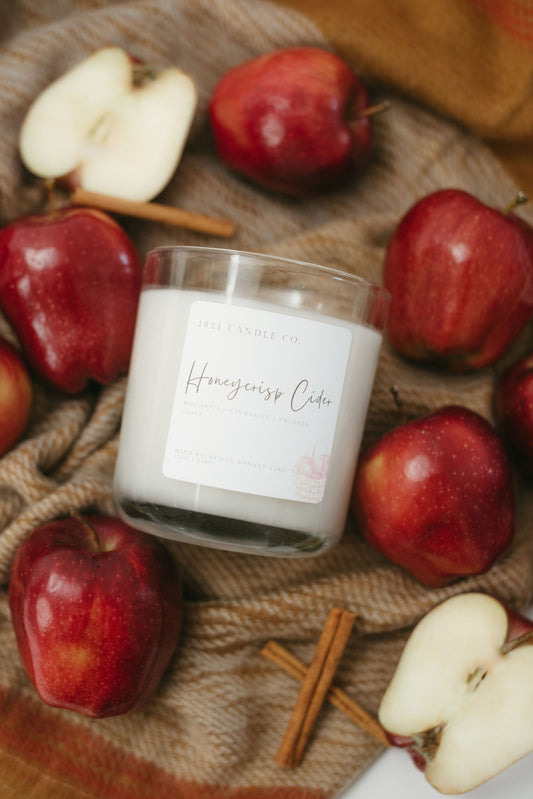Honeycrisp Cider Soy Blend Candle in clear reusable glass jar laying on a blanket surrounded by apples and cinnamon sticks. A cozy and warm fall candle.