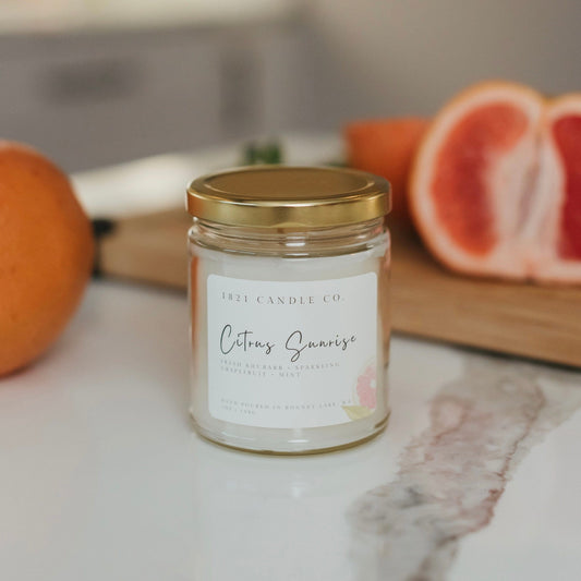 7oz Citrus Sunrise candle from 1821 Candle Co surrounded by sliced grapefruit and fresh greens - handcrafted home scent with fresh rhubarb, mint and grapefruit notes.