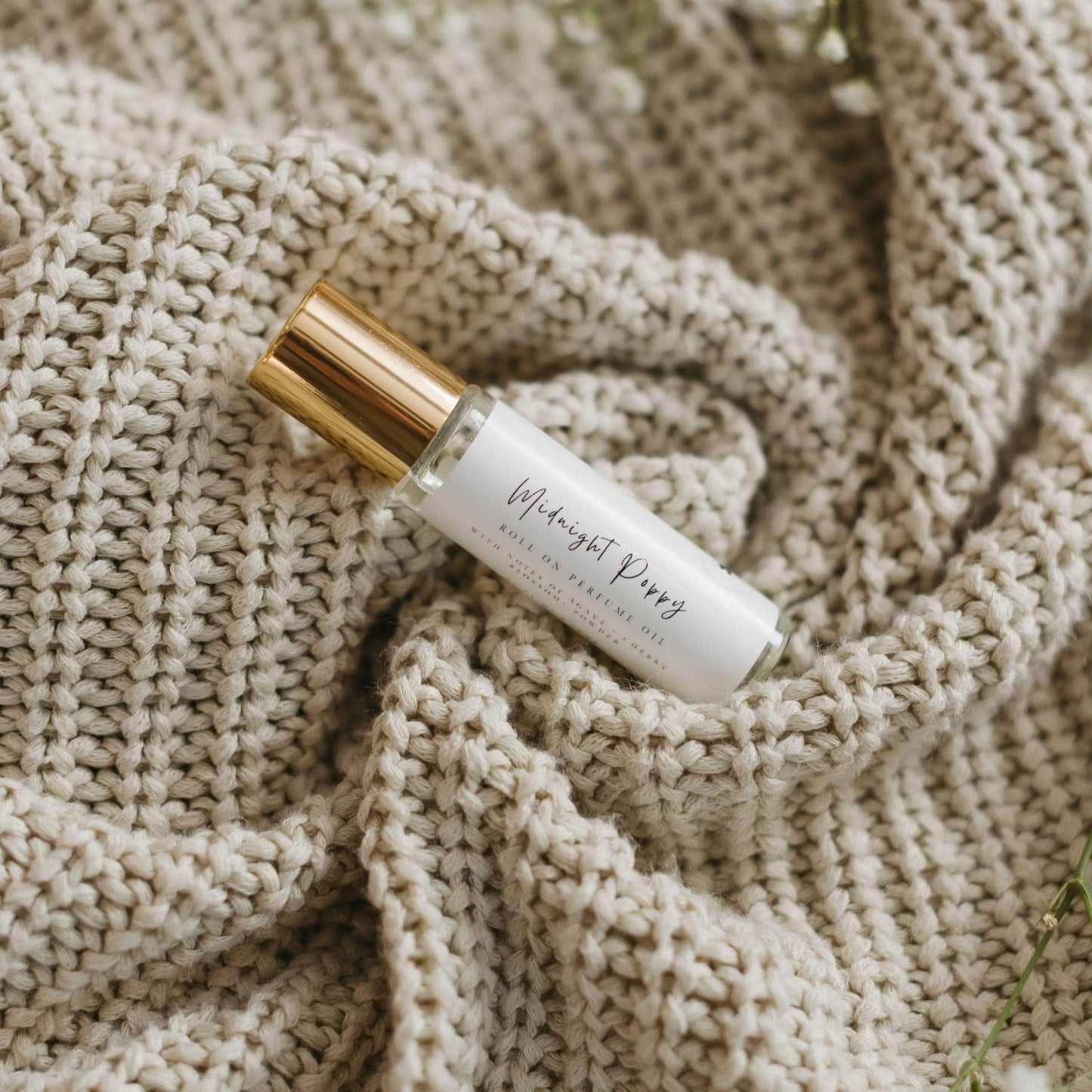 Midnight Poppy by 1821 Candle Co roller ball perfume features in a clear glass vial and gold lid with minimalist label laying on a cream colored knitted sweater with babys breath on the side. Notes of pear, agave, amber and musk.