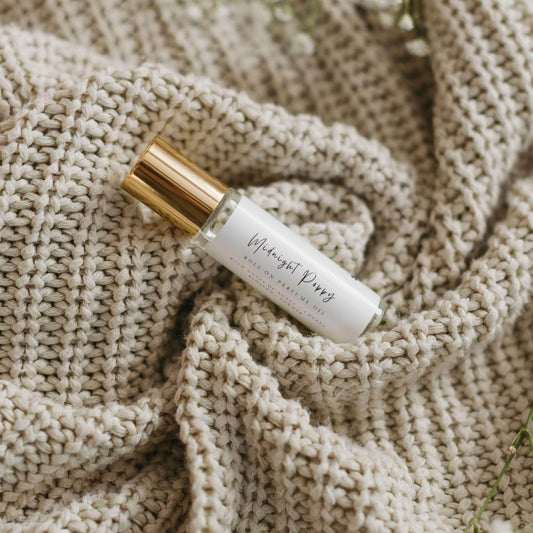Midnight Poppy by 1821 Candle Co roller ball perfume features in a clear glass vial and gold lid with minimalist label laying on a cream colored knitted sweater with babys breath on the side. Notes of pear, agave, amber and musk.