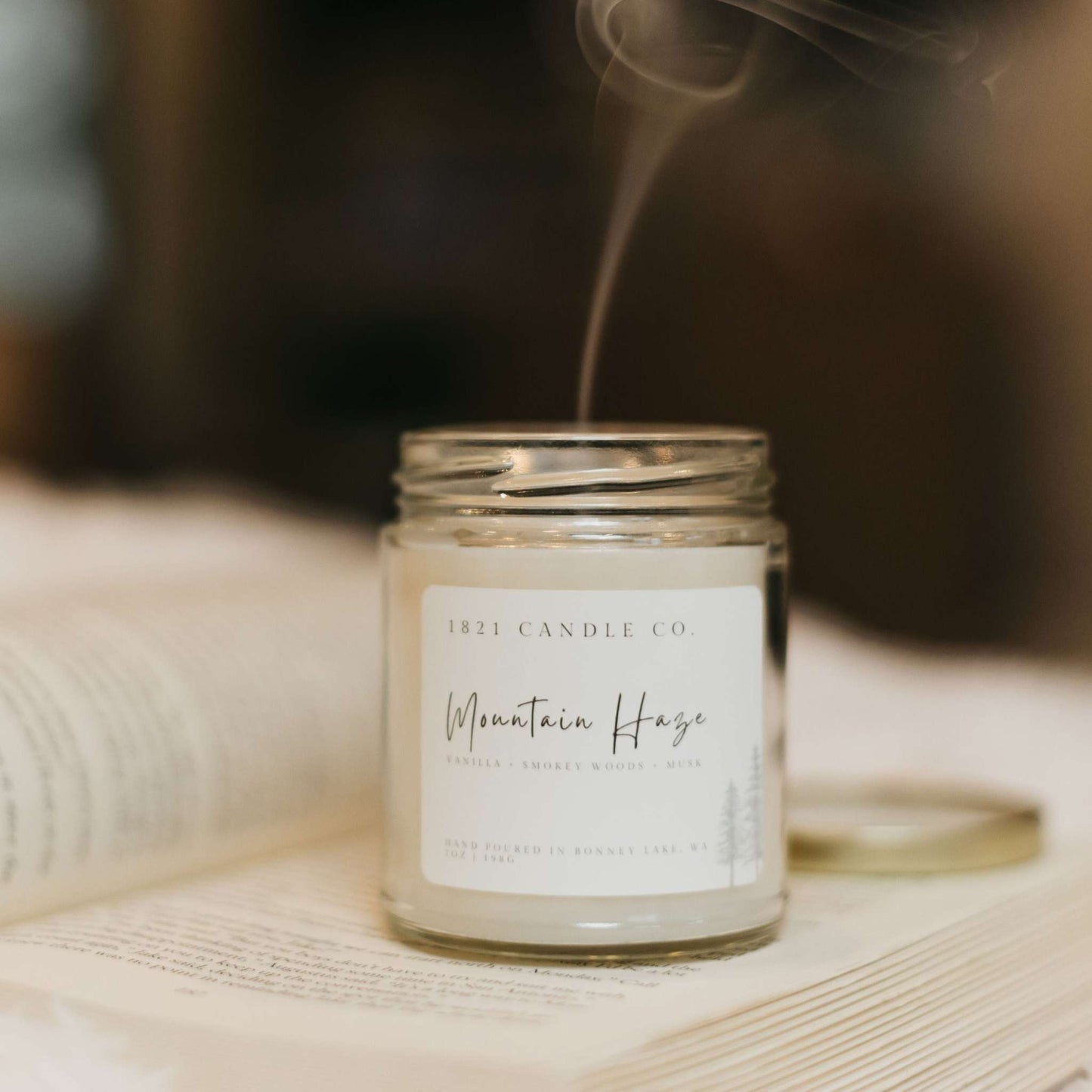 A premium soy blend candled called Mountain Haze by 1821 Candle Co. Minimalist farmhouse style candle sitting on top of an open book and candle was just lit and blown up. Small stream of smoke coming up from candle wick.