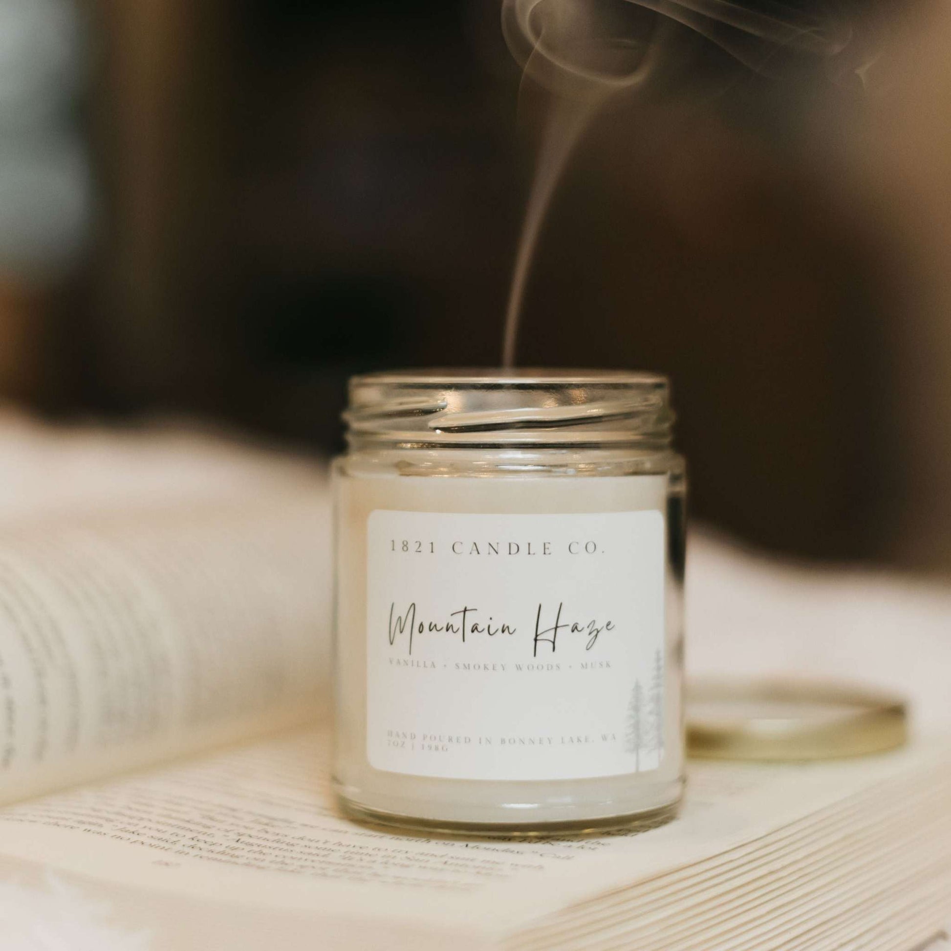 A premium soy blend candled called Mountain Haze by 1821 Candle Co. Minimalist farmhouse style candle sitting on top of an open book and candle was just lit and blown up. Small stream of smoke coming up from candle wick.
