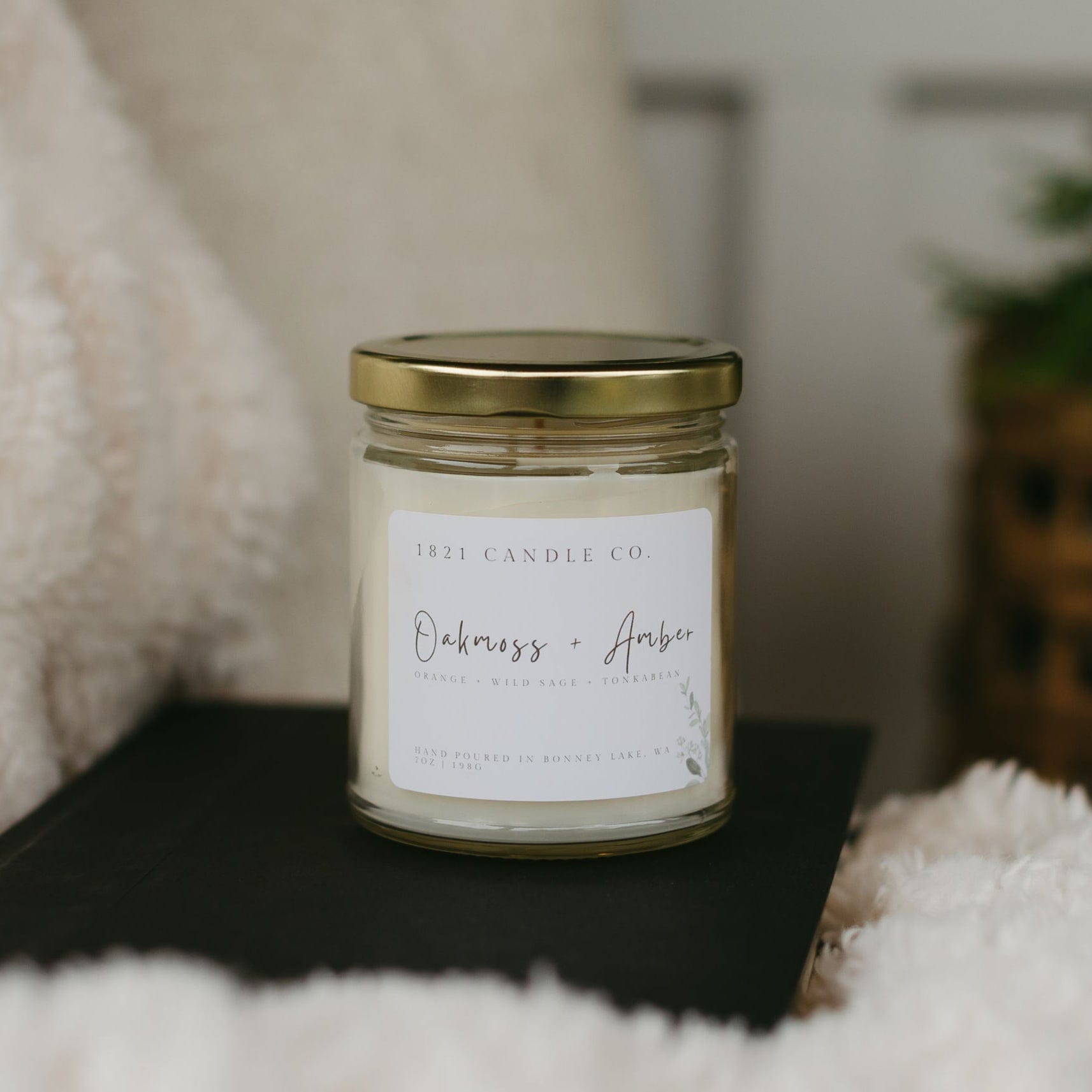 Close up of Oakmoss Amber soy candle on a black book with a plush white blanket - a fresh home fragrance.
