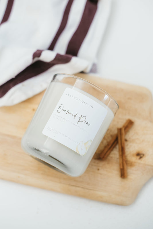 Orchard Pear Candle with notes of pear, cinnamon, honey, nutmeg laying on a cutting board with cinnamon sticks, sprinkled with cinnamon and a kitchen towel.