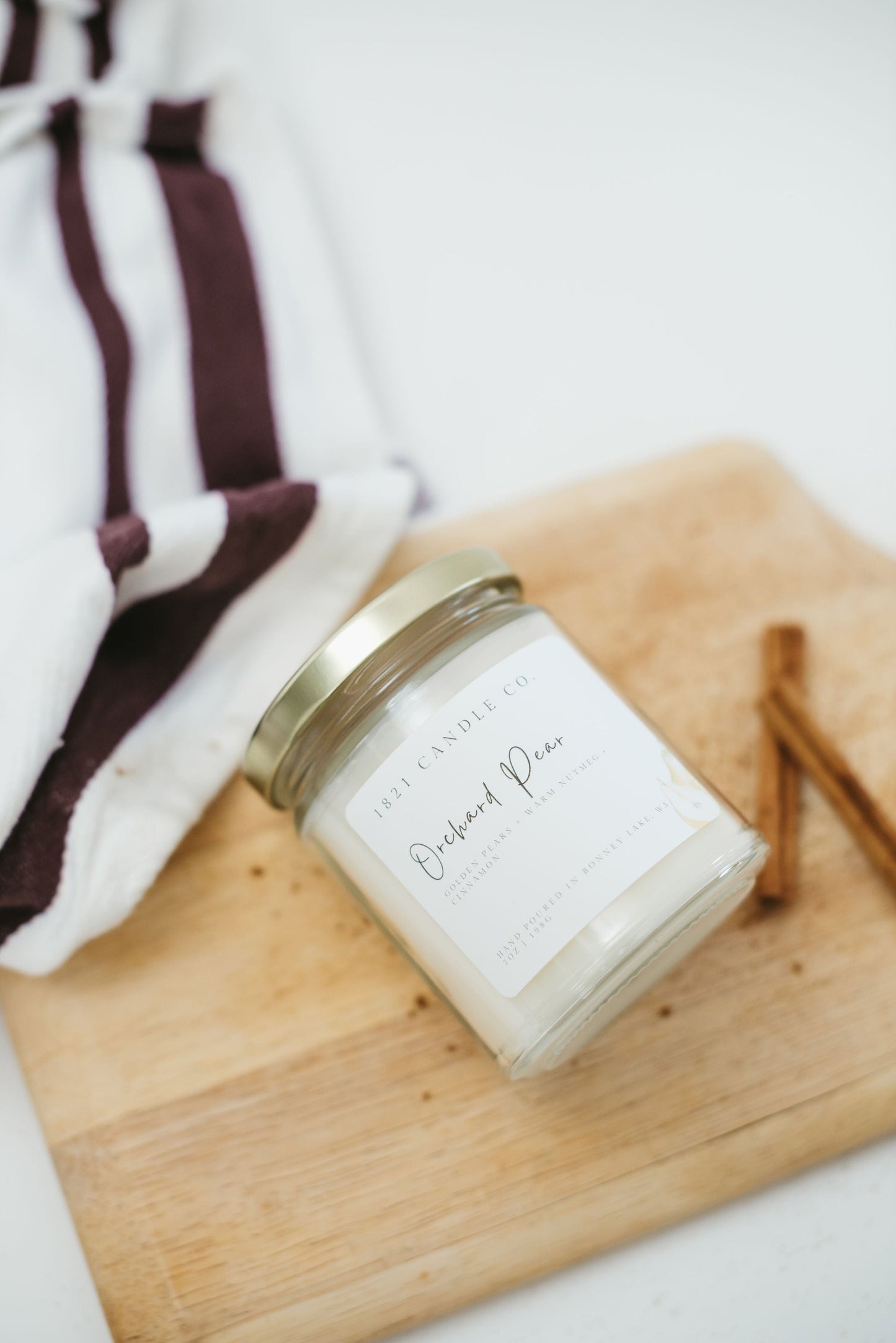 Orchard Pear Candle by 1821 Candle Co laying on it's side with a cutting board underneath and cinnamon sticks on the side.