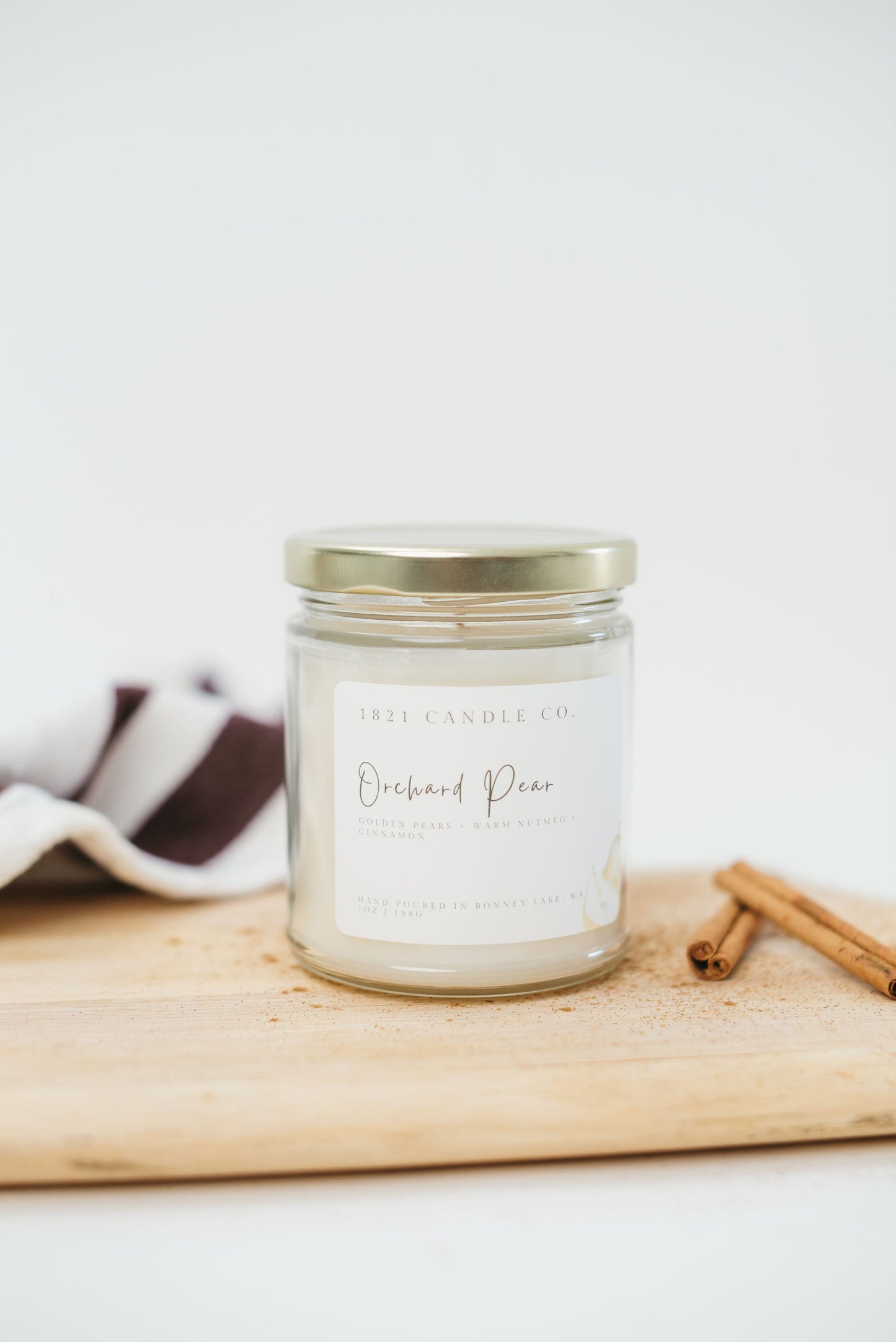 Orchard Pear Soy Blend Candle displayed on top of a cutting board with a white background and a kitchen towel and cinnamon sticks on the side.