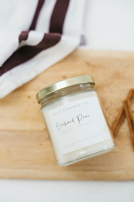 Orchard Pear soy blend fall candle laying on side with a cutting board and cinnamon sticks with a kitchen towel.