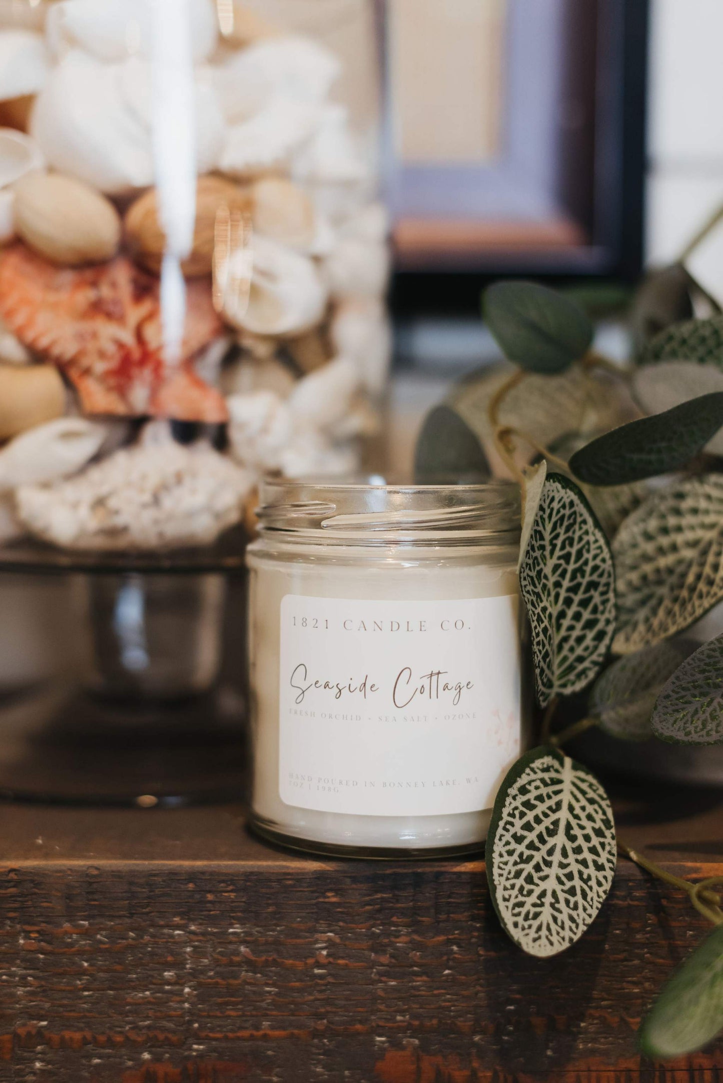 A hand poured soy candle called Seaside Cottage in a glass jar sitting on a fireplace ledge surrounded by soft neutral toned decor and a vase full of seashells.