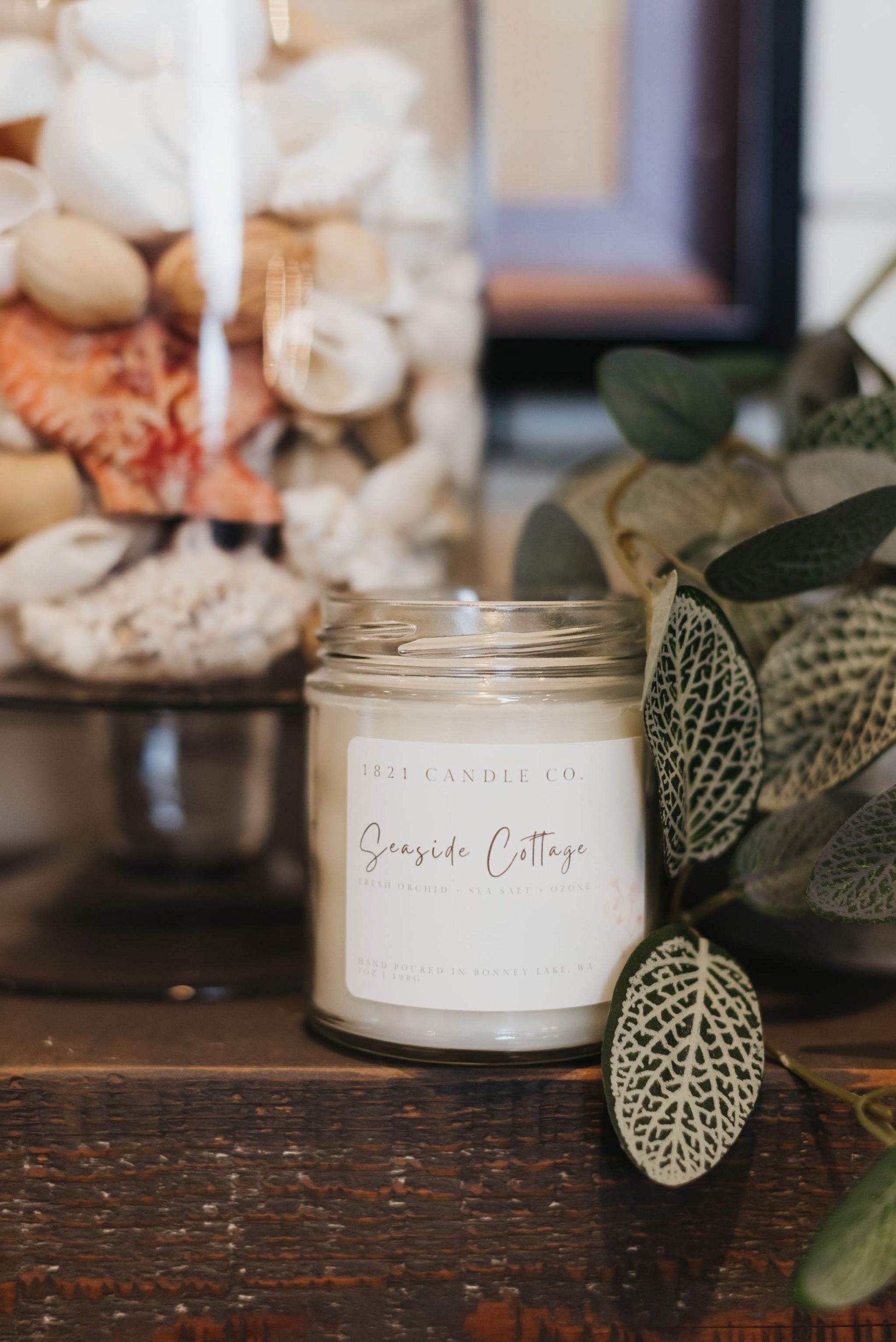 A hand poured soy candle called Seaside Cottage in a glass jar sitting on a fireplace ledge surrounded by soft neutral toned decor and a vase full of seashells.