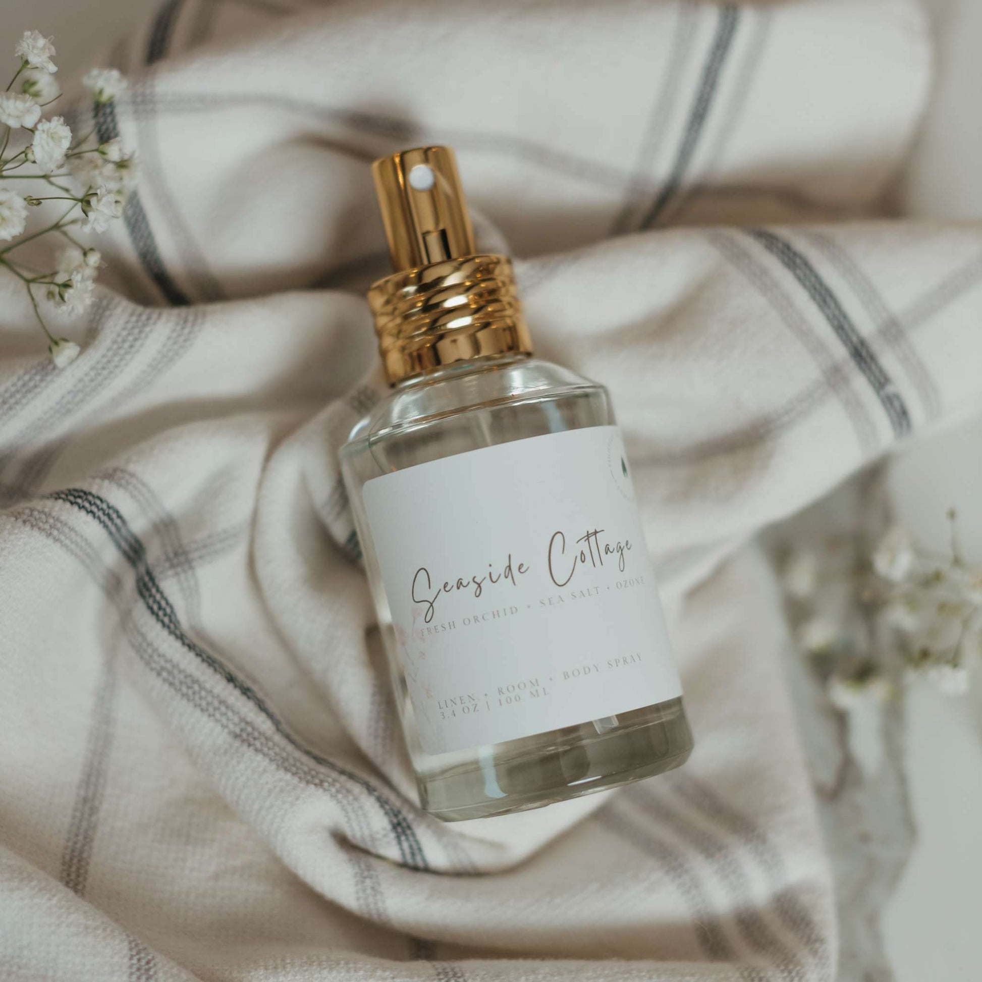 Seaside Cottage linen, room and body spray in a glass bottle with a gold fine mist nozzle, resting on a soft, neutral plaid fabric with delicate white florals. A fresh coastal inspired fragrance featuring fresh orchid, sea salt and ozone.