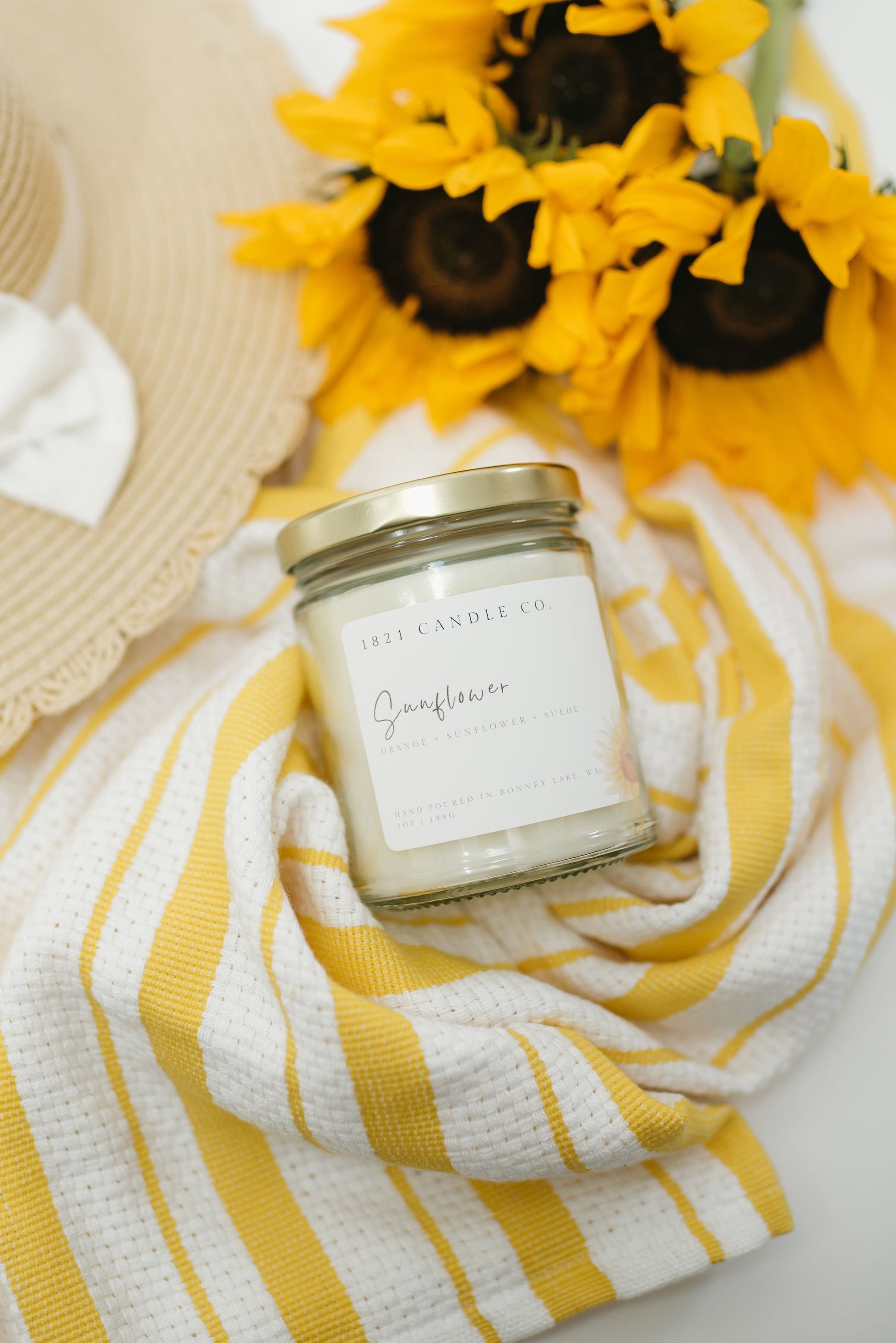 1821 Candle Co sunflower candle styled on a yellow striped towel with sunflowers and a sun hat - summer inspired candle gift for cozy home lovers.