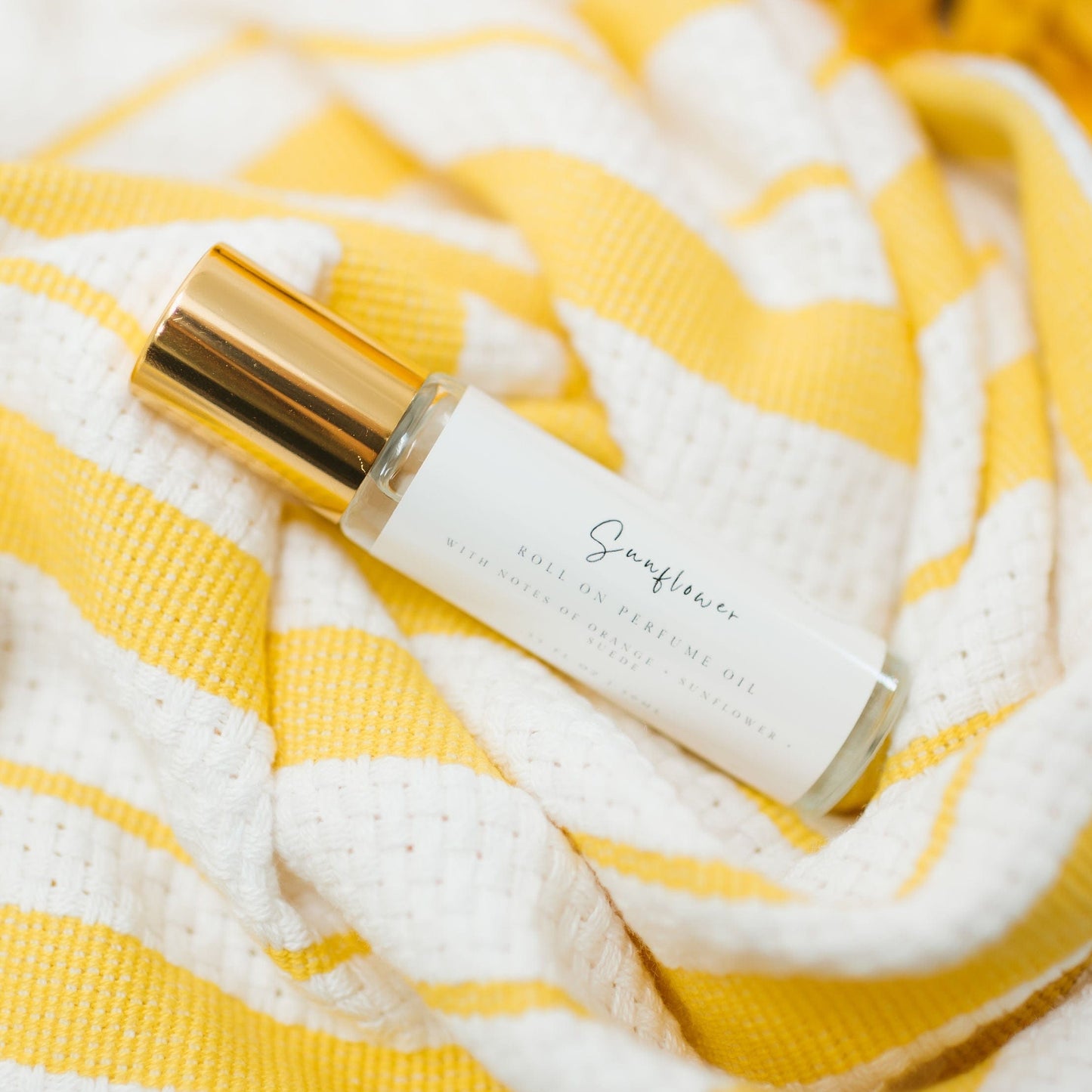 Sunflower Roller Ball Perfume made with Coconut Oil and skin safe fragrance oil. A classic gold capped bottle with a minimalist farmhouse style label laying on a bright stripped yellow kitchen towel.