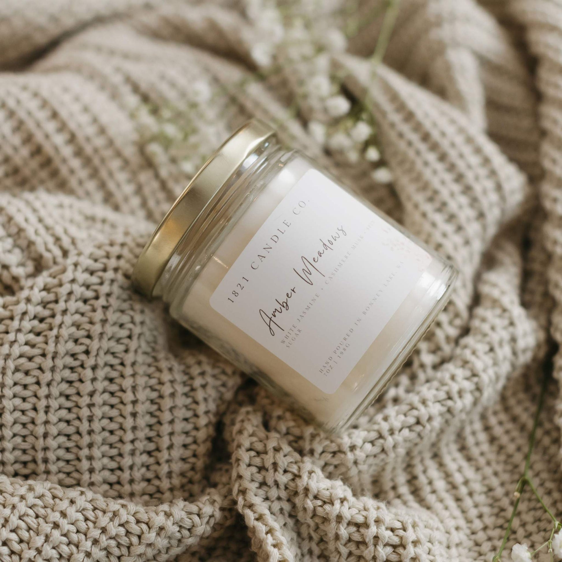 Amber Meadows by 1821 Candle Co made in small batches, laying on it's side on top of a cream colored knitted cardigan with baby breath on next to it. Notes of spun sugar, cashmere musk, jasmine.