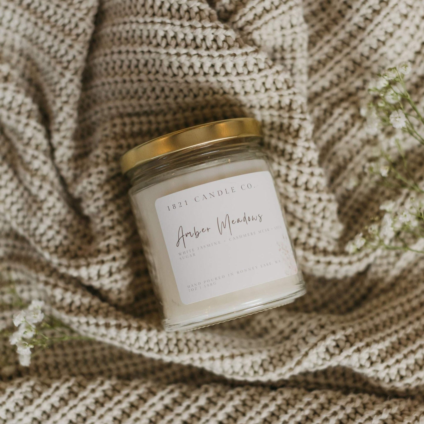A small batch soy blend candle called Amber Meadows by 1821 Candle Co laying on it's side on top of a cream knitted cardigan with baby breath laying next to it.