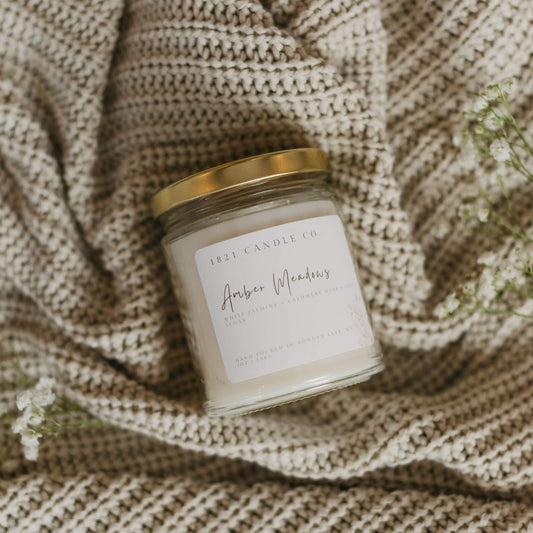 A small batch soy blend candle called Amber Meadows by 1821 Candle Co laying on it's side on top of a cream knitted cardigan with baby breath laying next to it.
