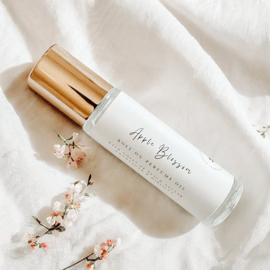 Glass bottle roller perfume with gold lid laying on a white linen sheet surrounded by apple blossom flowers.