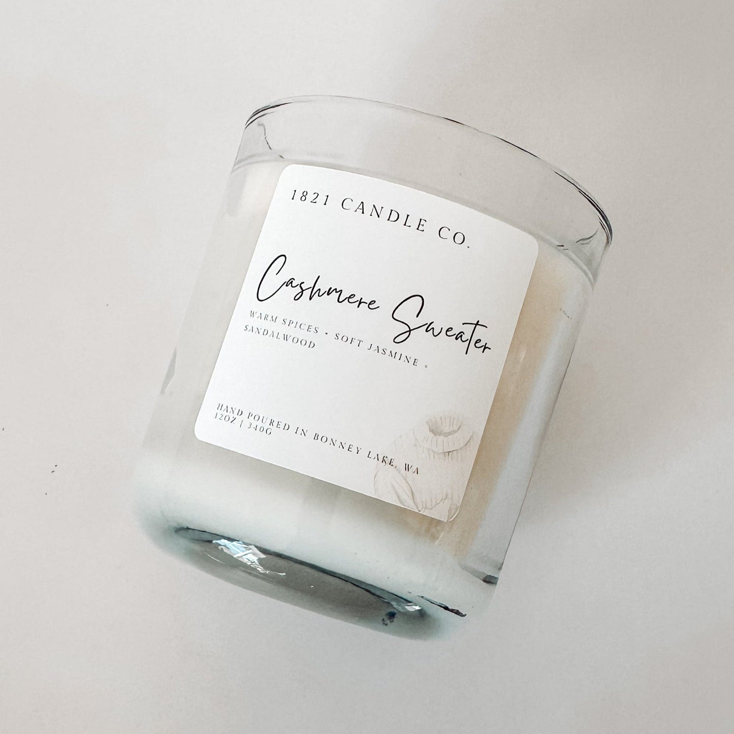 1821 Candle Co. Cashmere Sweater candle lying on a white surface, showcasing its clean minimalist label and cozy scent profile of soft jasmine and spices.