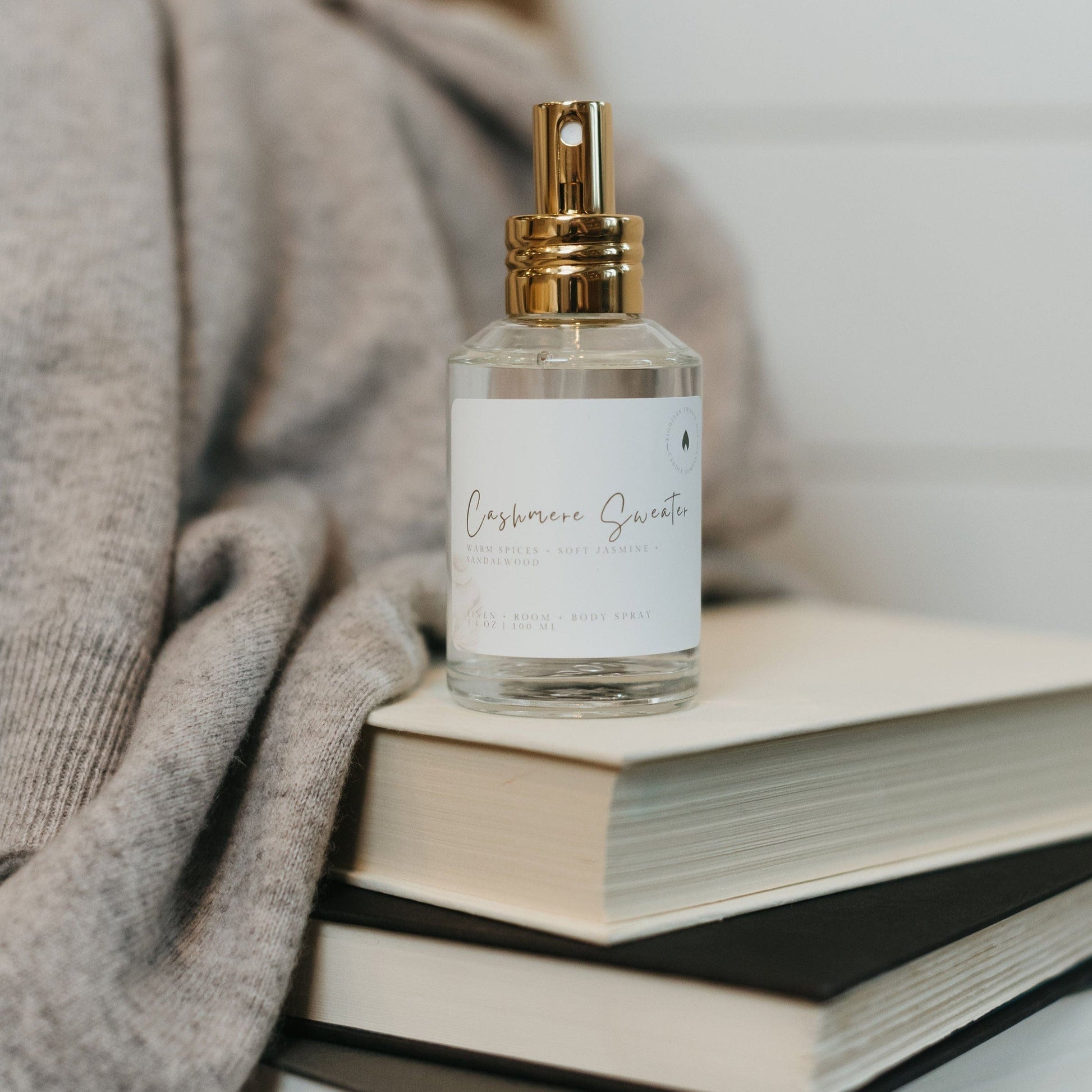 Cashmere Sweater room spray placed on a stack of books with a soft gray cashmere sweater, creating a cozy, inviting atmosphere. Warm vanilla, sandalwood and amber scent.