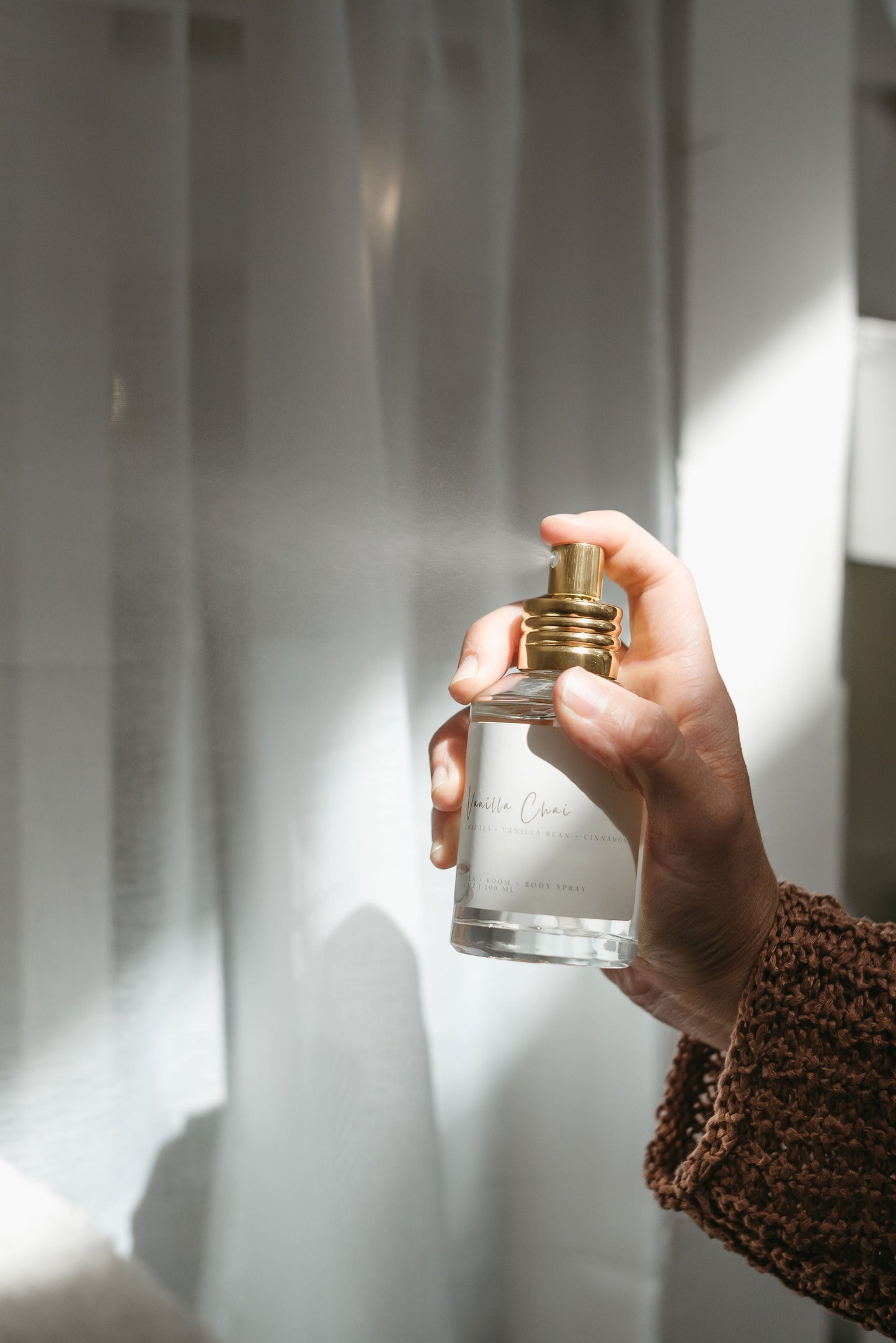 Hand spraying fall air freshener called Vanilla Chai against a white sheet curtain.