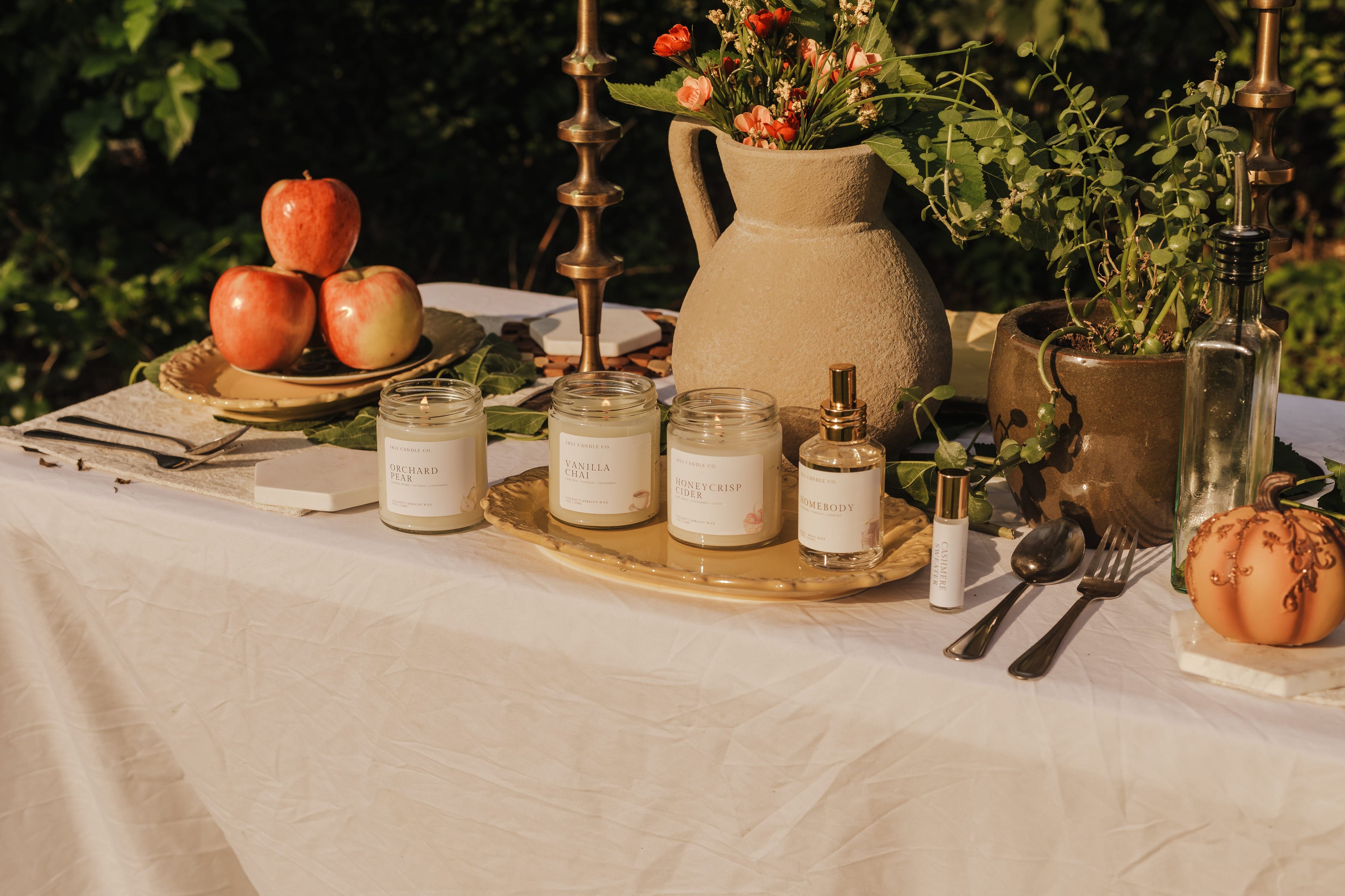 A styled outdoor table setting with several glass jar candles on a gold tray, alongside apples, a ceramic pitcher with flowers, greenery, cutlery, and a small pumpkin, all arranged on a white tablecloth. Photography by Tiffany Carr Collective