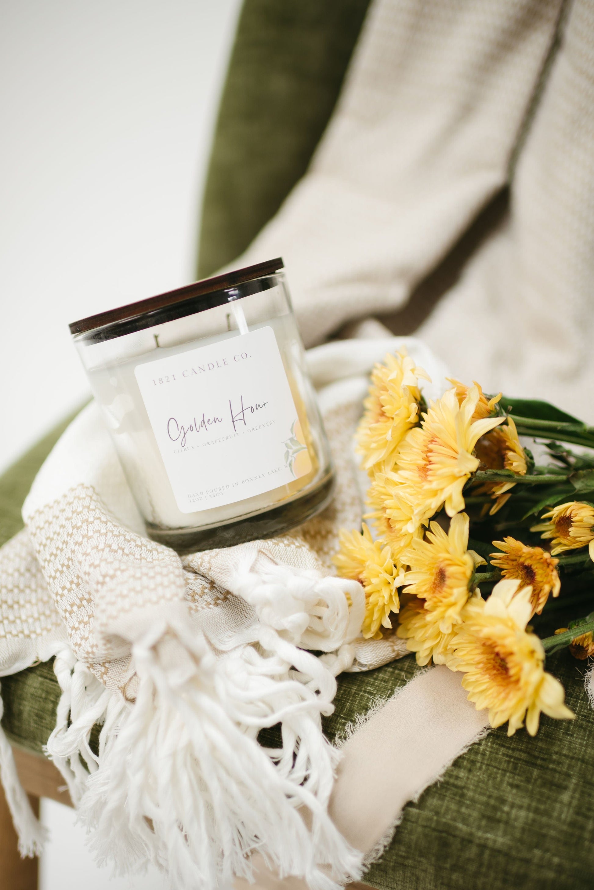 Golden Hour 12oz candle co styled with yellow daisies and neutral blankets on a green chair - an inviting citrus home fragrance for slow living spaces.