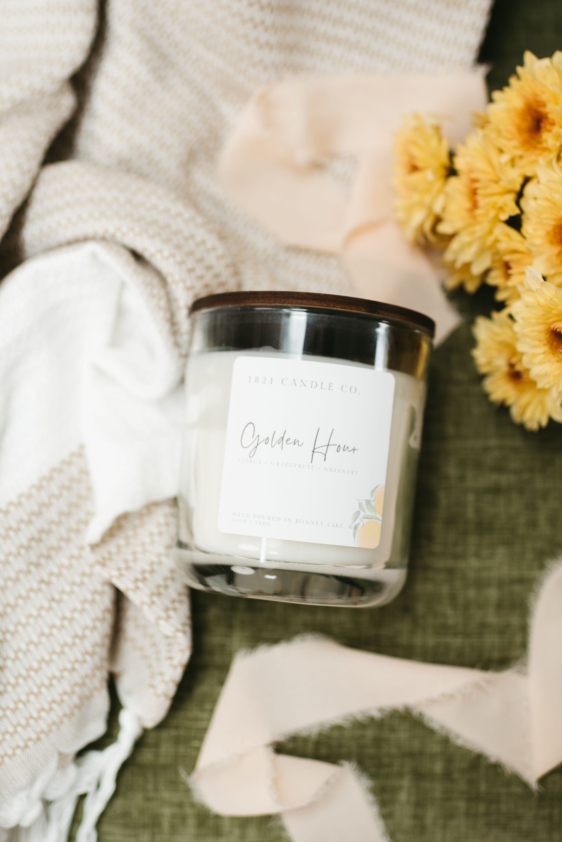 12oz Golden Hour Candle in a glass jar with wooden lid, photographed with yellow flowers and soft blankets - perfect non toxic citrus candle for cozy home decor lovers.