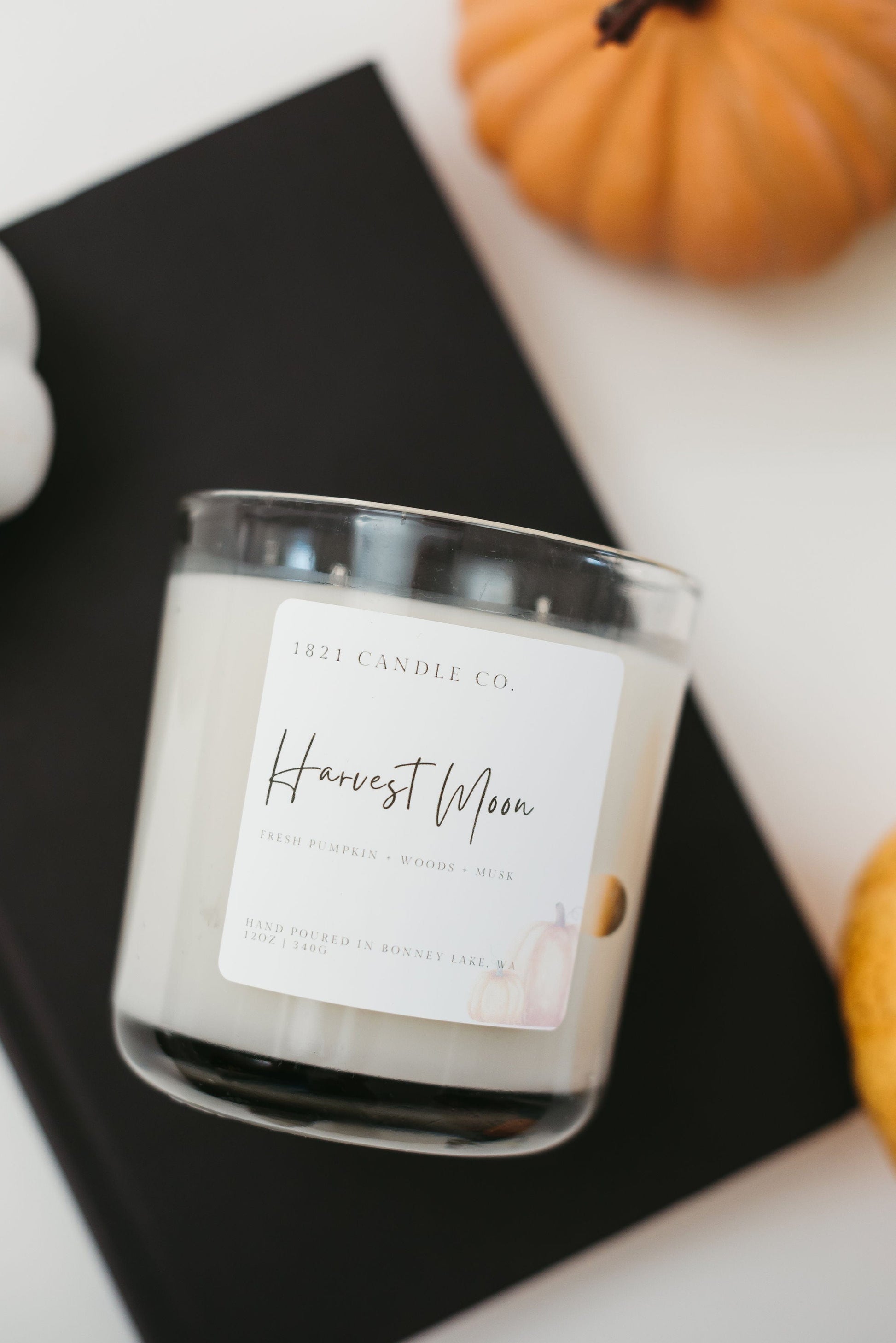 Harvest Moon soy blend candle in 12oz clear jar with notes of fresh pumpkin, patchouli, spiced apple, leaves, musk laying on top of a book with pumpkins placed next to it.