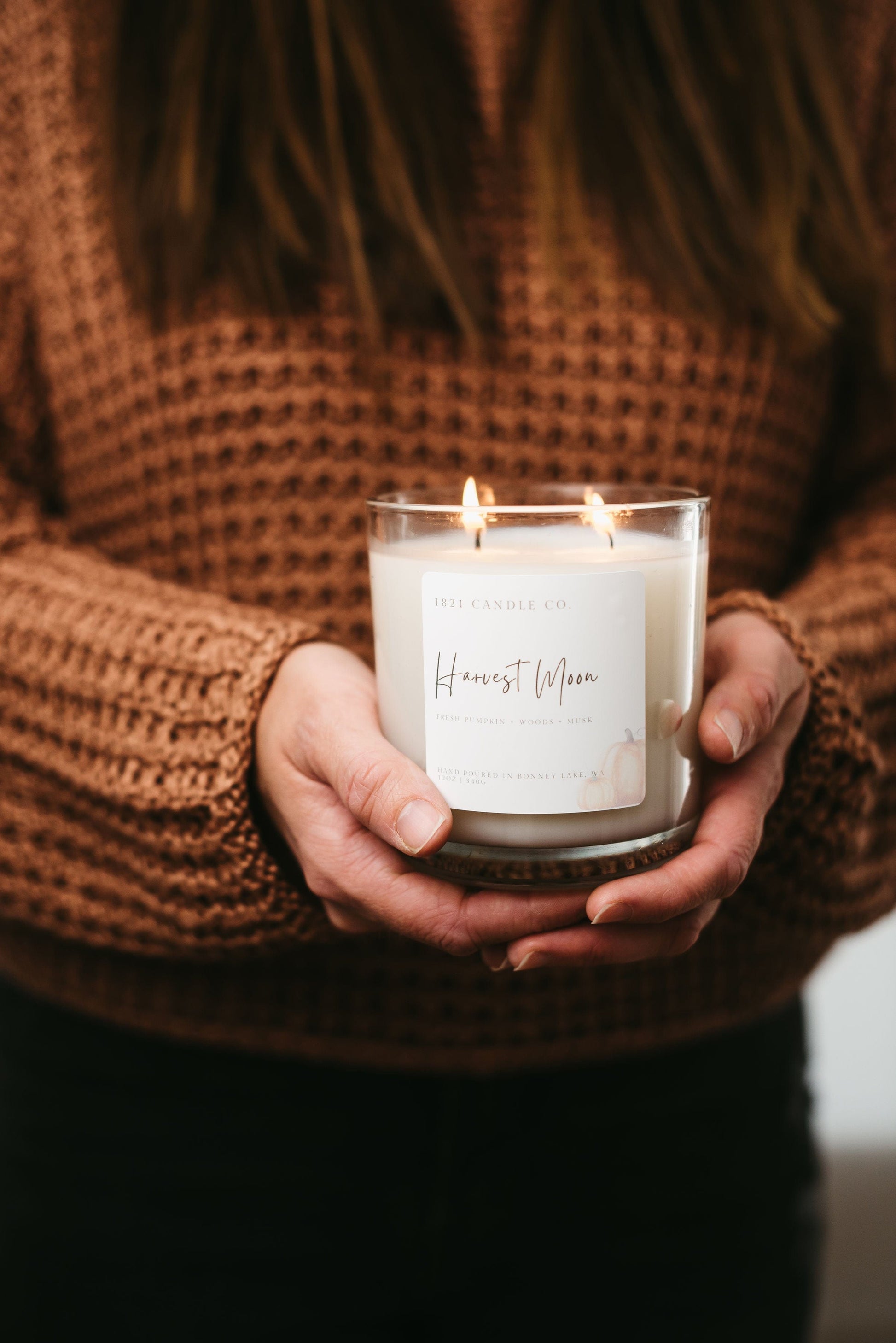 Person holding a lit candle in a glass holder with 'Harvest Moon' text, wearing a brown textured sweater.