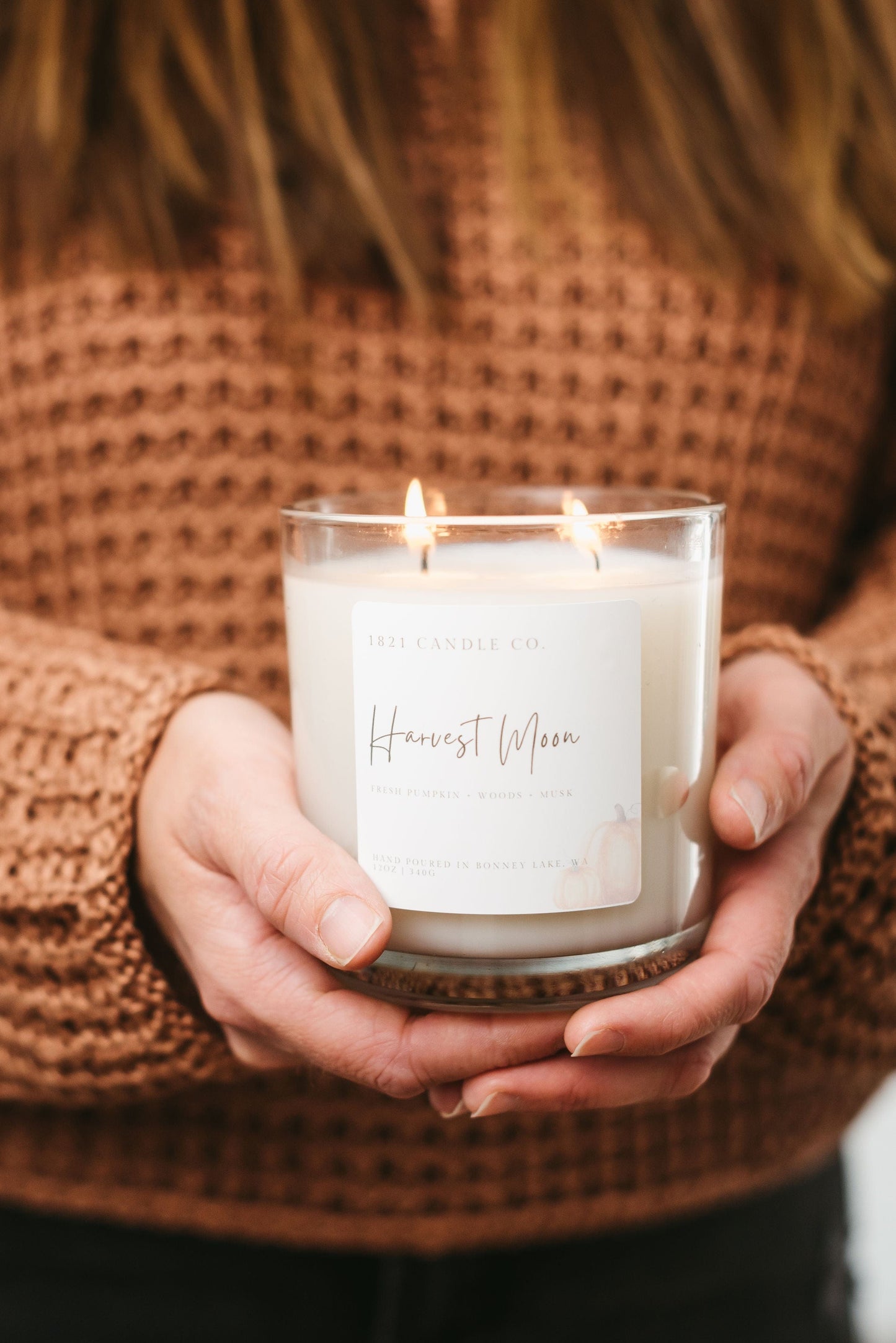 Person holding a lit candle labeled 'Harvest Moon' against an orange sweater