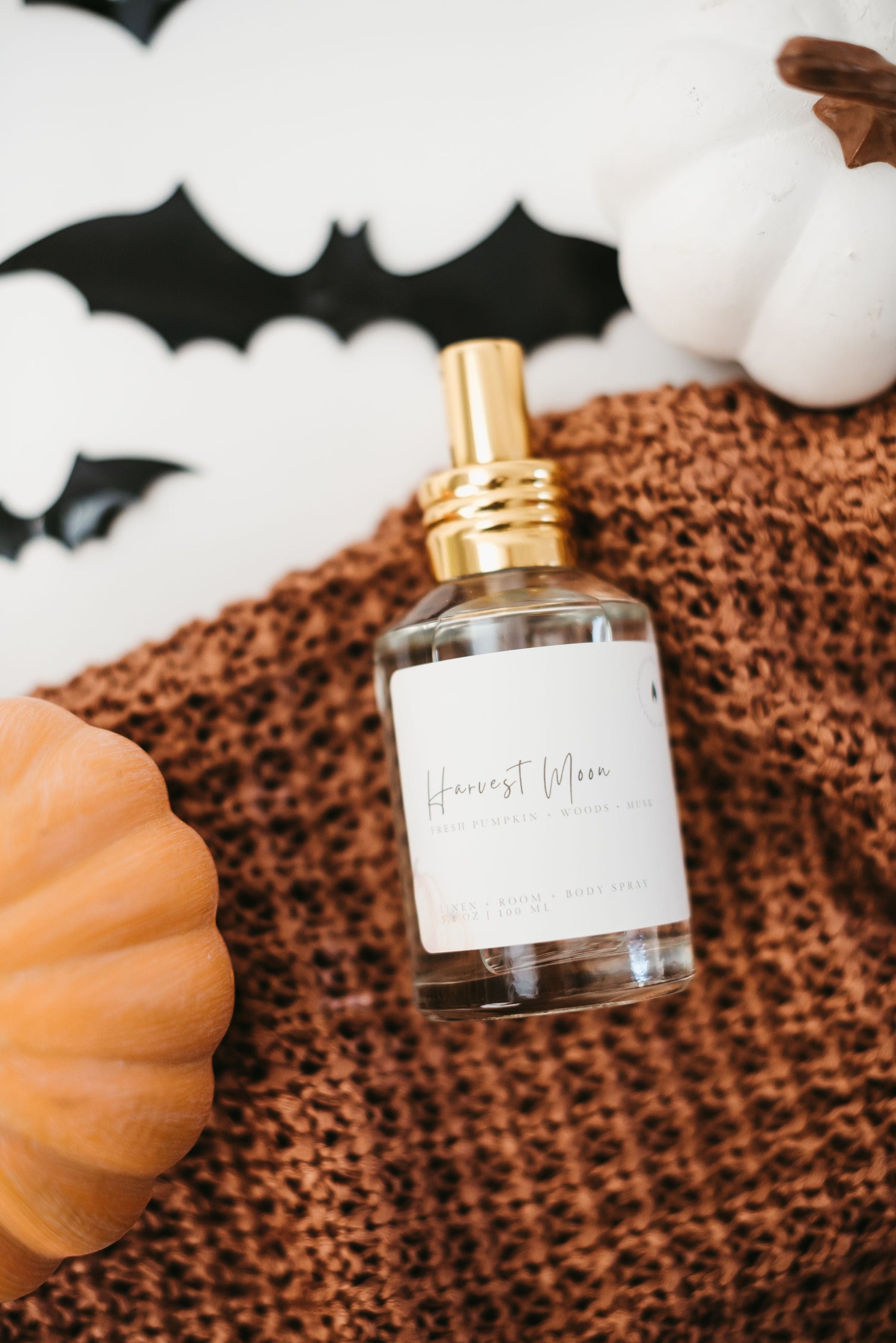 Harvest Moon Room, Linen, Body Spray in a clear glass bottle and gold lid laying on a orange sweater with fall pumpkins in the background.