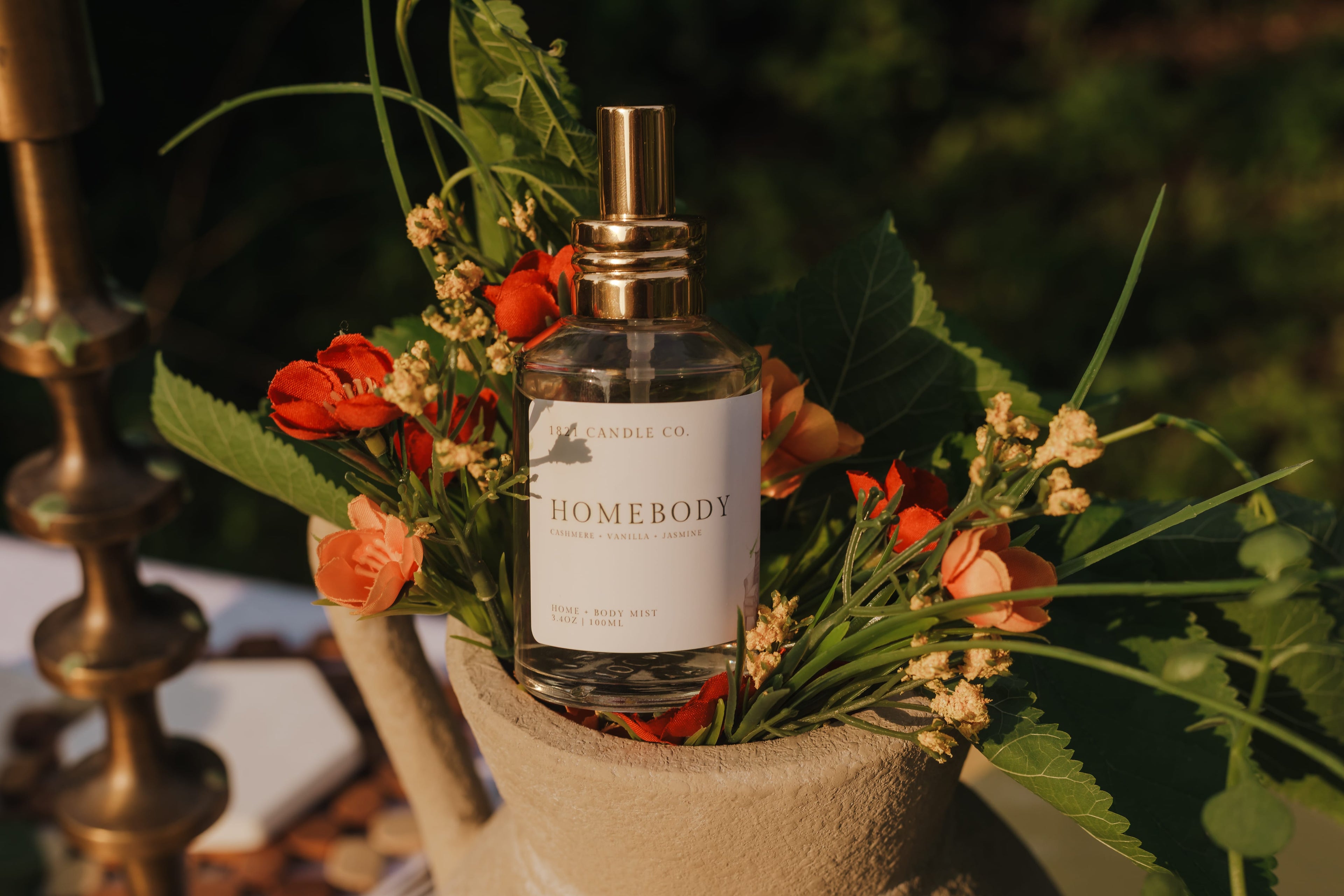 A bottle of Homebody scented room spray, body spray, linen spray by 1821Candle Co. with a gold spray top, nestled in a rustic arrangement of green leaves, orange flowers, and dried botanicals in a beige pot. Photography by Tiffany Carr Collective.