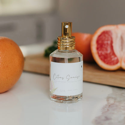 Clear glass bottom of Citrus Sunrise linen and room spray with a gold sprayer, displayed next to fresh grapefruit and mint on a marble countertop - non toxic home fragrance for everyday freshness.
