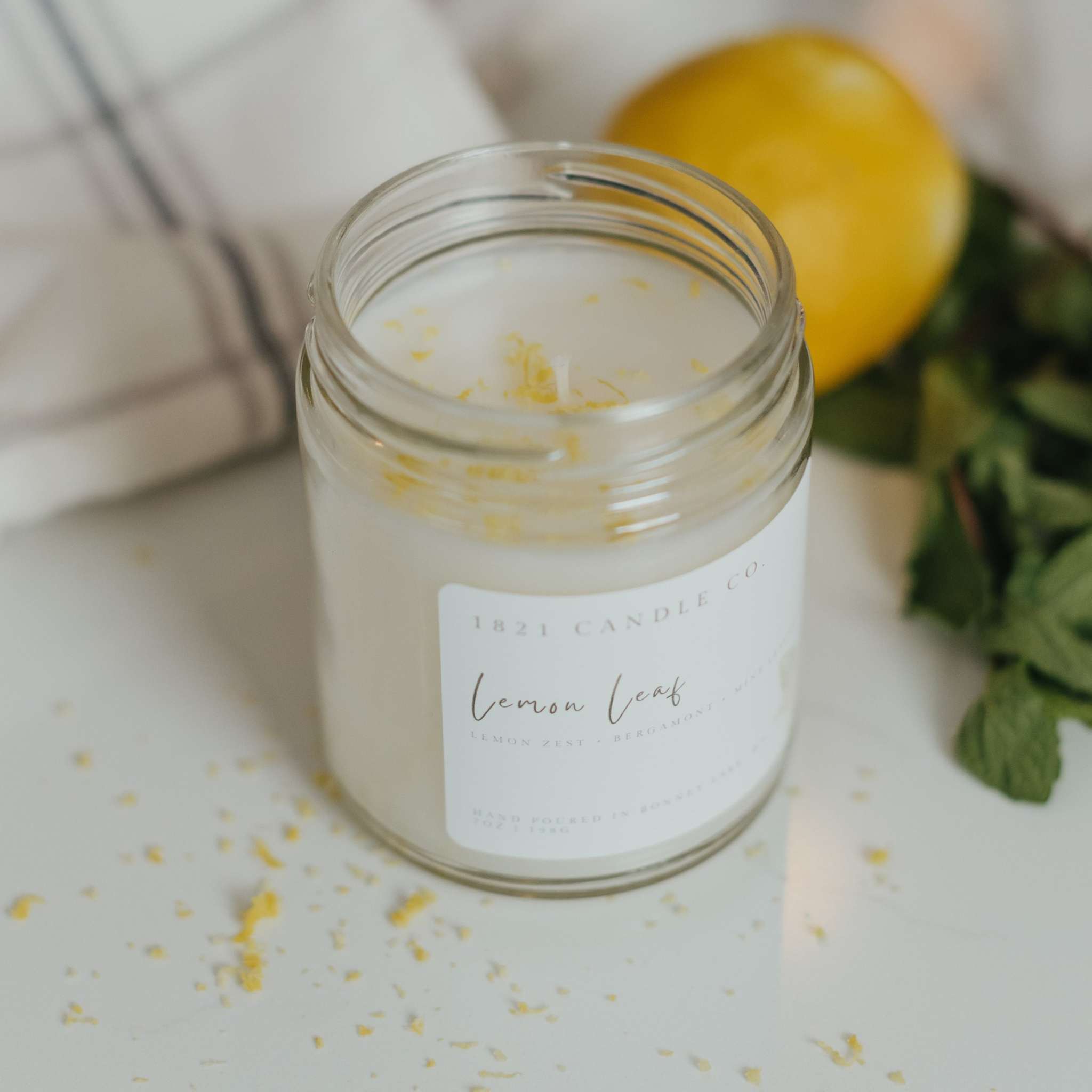 Lemon Leaf Candle by 1821 Candle Co on a white marble kitchen counter with fresh lemon and mint leaves in the background laying next to a neutral plaid kitchen towel. Freshly grated lemon zest lays in front of the candle.