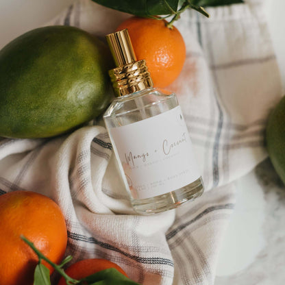 Mango Coconut Room Spray in a glass bottle with a gold sprayer, styled with fresh mangoes and oranges on a striped linen towel. Tropical home fragrance for a fresh uplifting scent.