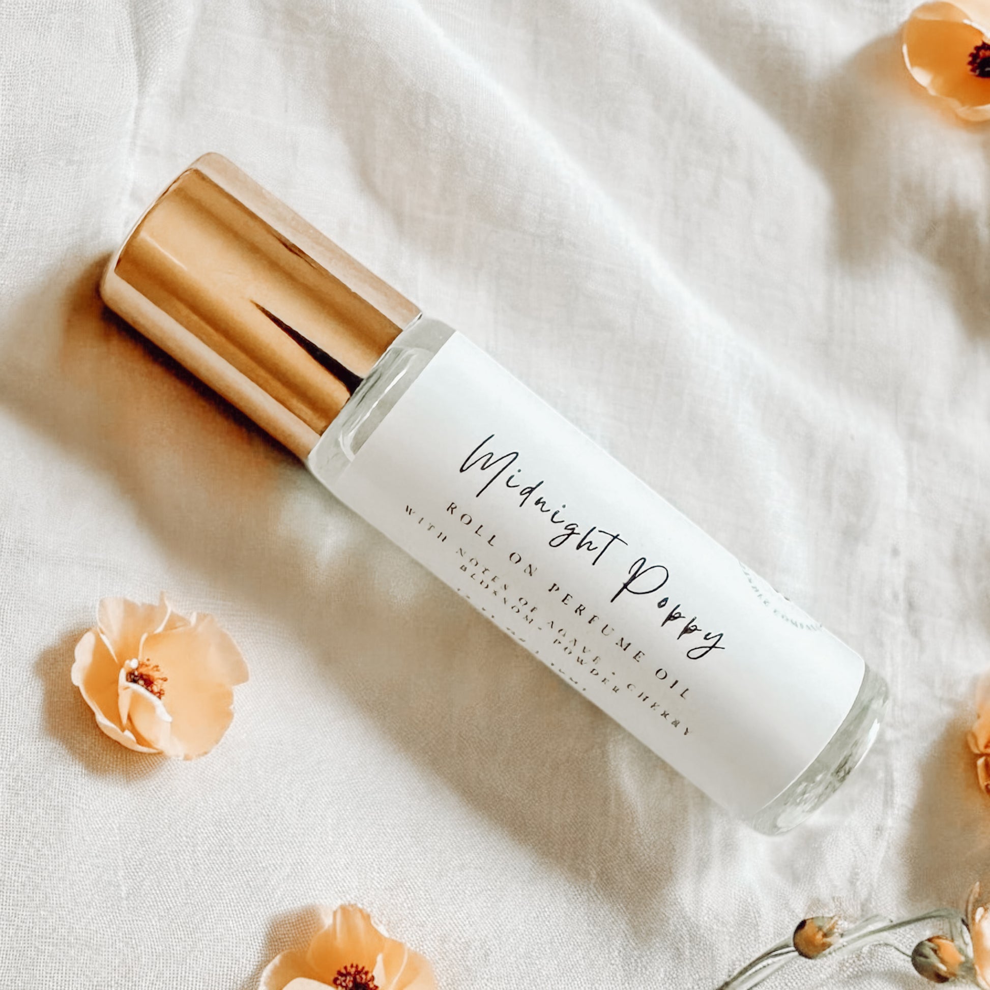 Glass bottle roller perfume with gold lid laying on white linens surrounded by yellow poppy flowers.