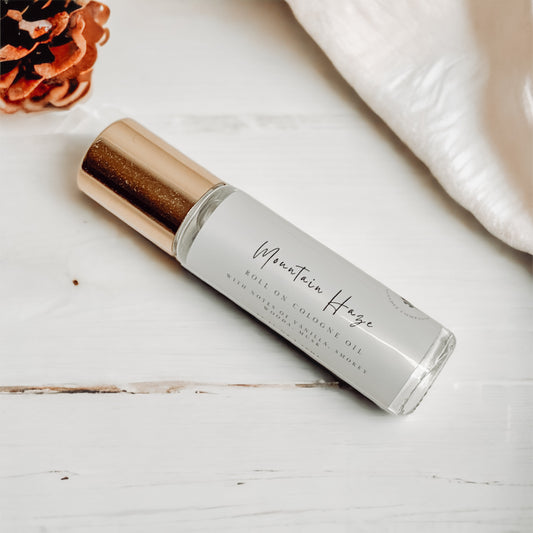 Glass bottle cologne roller with gold cap laying on a white wooden surface with a pine cone and fabric in the top corners.