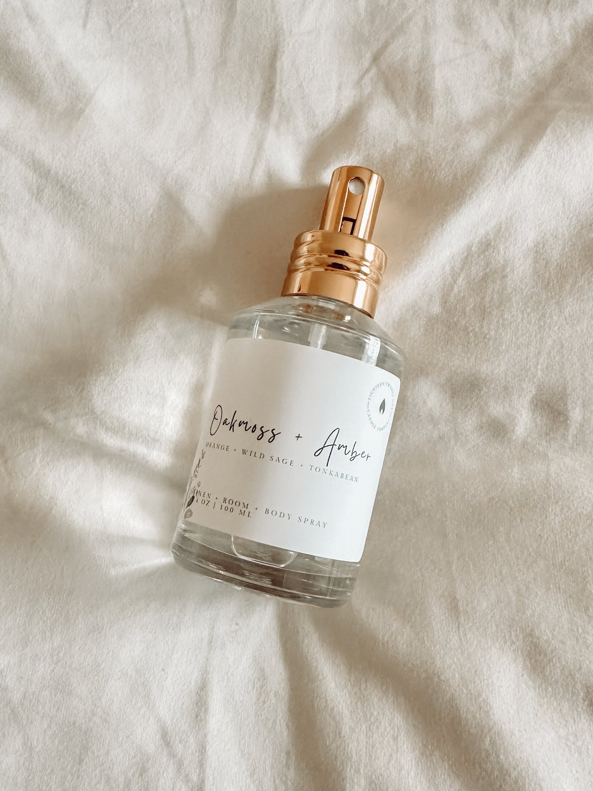 A clear spray bottle with gold lid called Oakmoss + Amber laying on side on top of a white sheet.