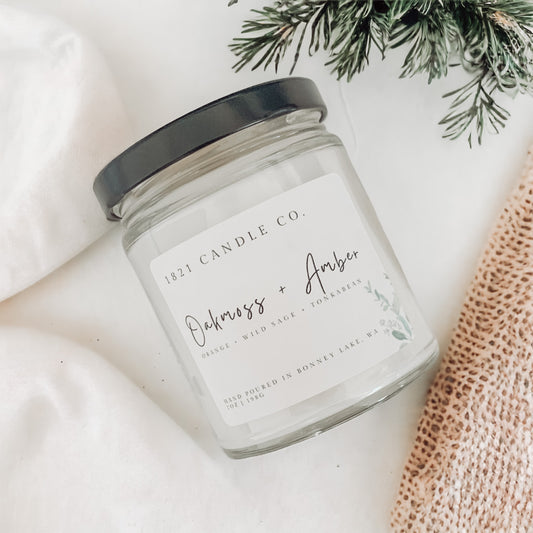 clear glass jar candle with black lid laying on a white background with brown textured linen and greenery on the side