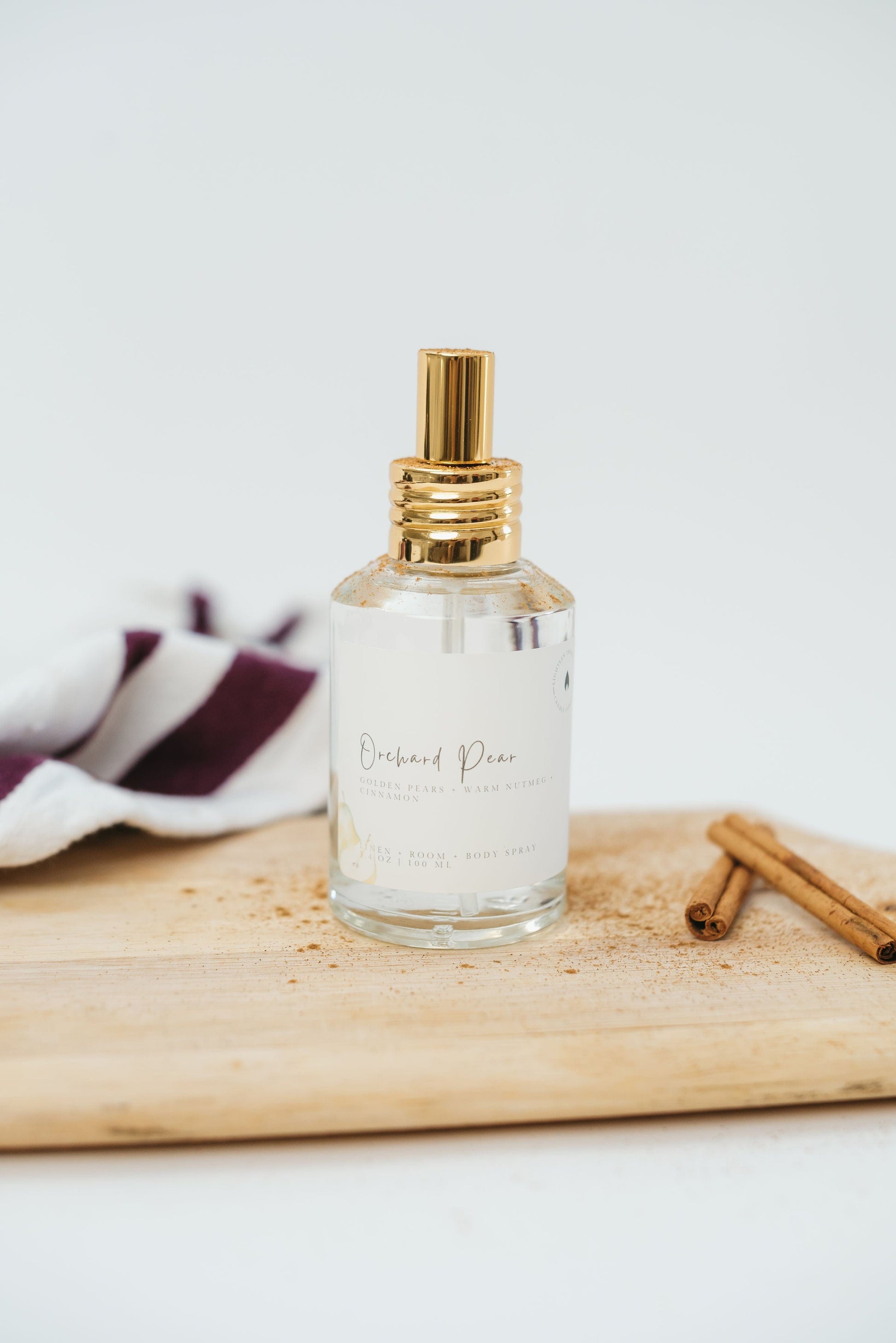 Perfume bottle with gold cap on a wooden board with cinnamon sticks and a striped towel in the background.