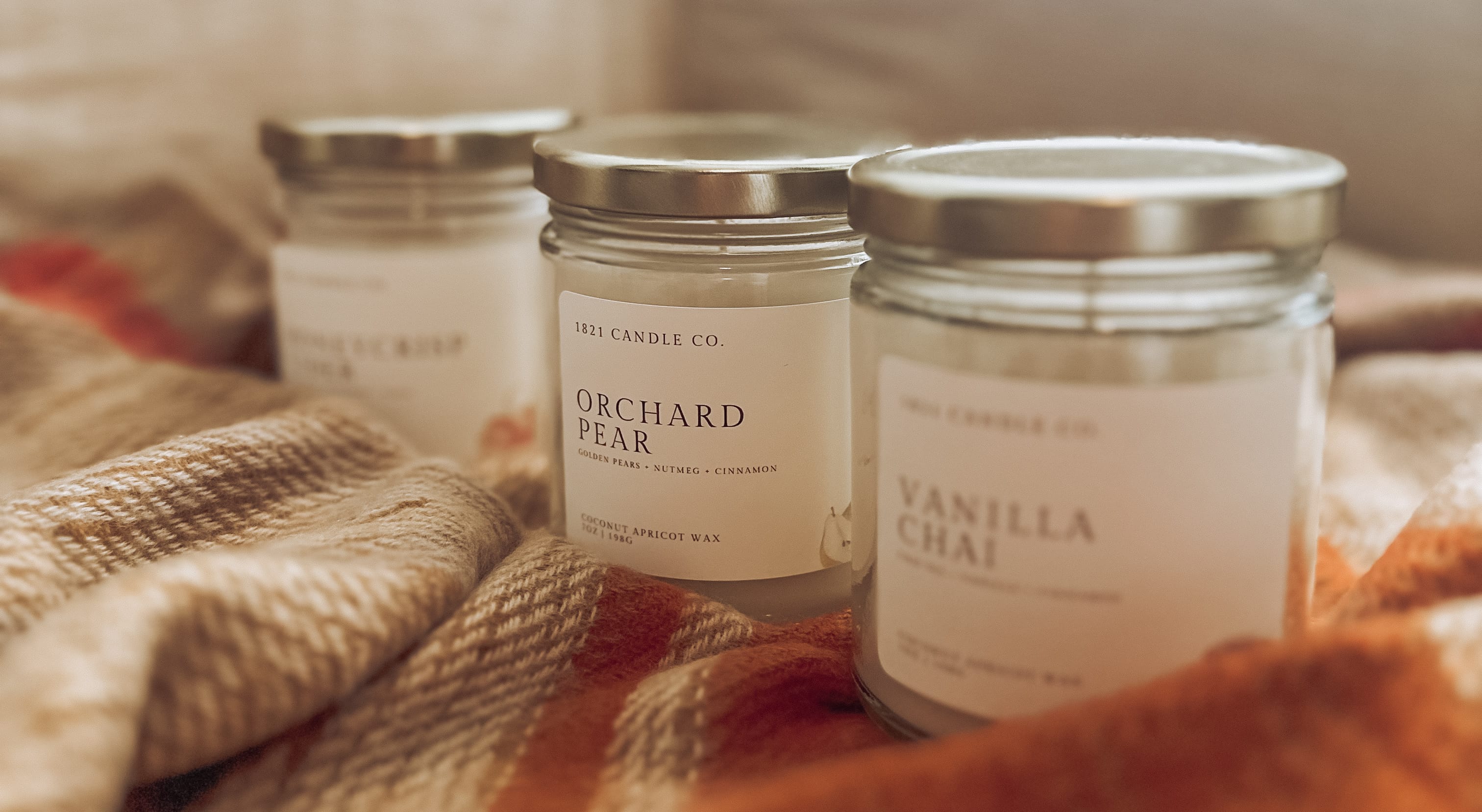 Three glass jar candles with gold lids are arranged on a cozy beige and rust plaid scarf. The front candle is labeled Vanilla Chai, with two more candles behind it, one labeled Orchard Pear and another partially visible in the back labeled Honeycrisp Cider. Small batch candles, handmade in Washington State.