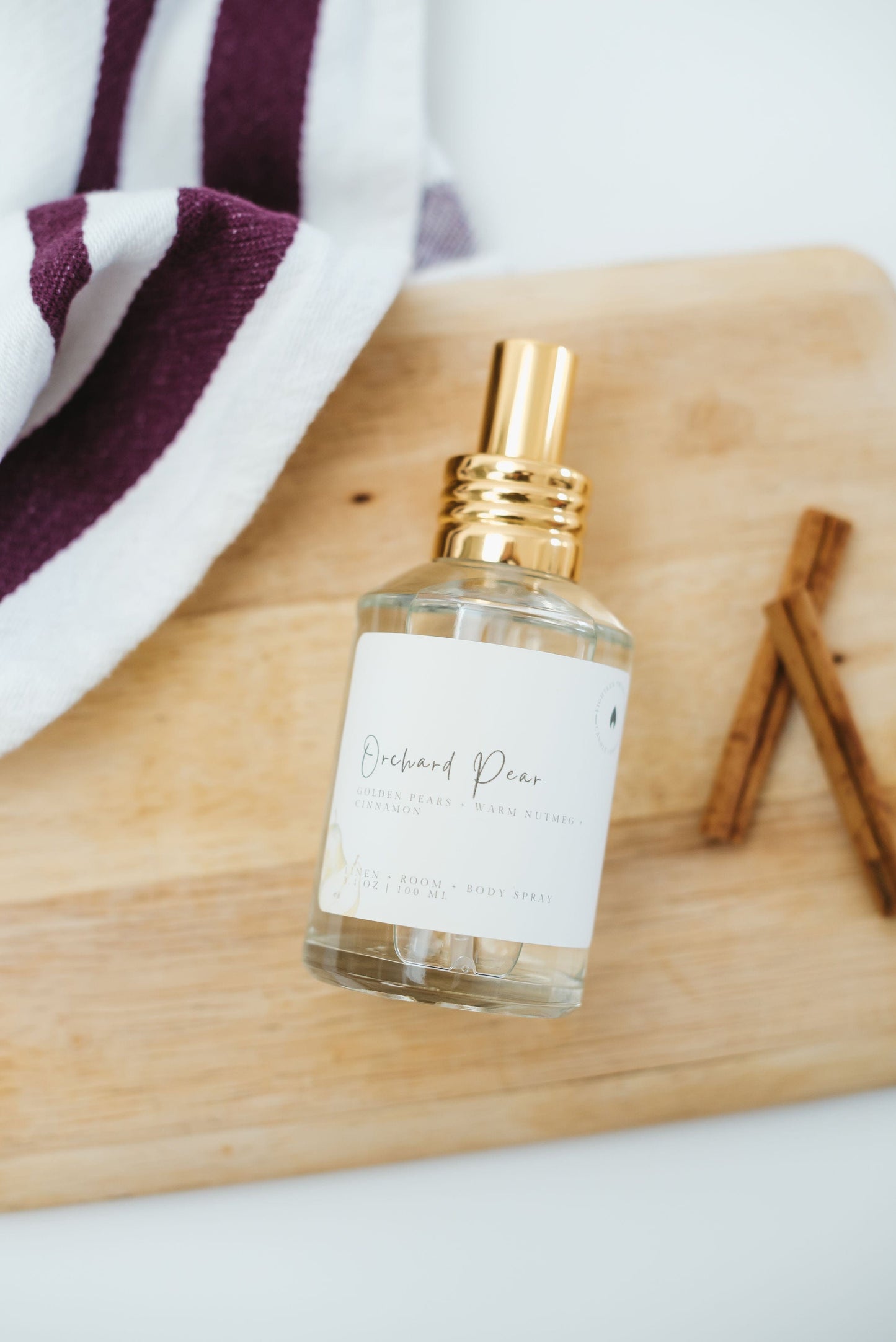Bottle of Orchard Pear linen, room, body spray  on a wooden surface with cinnamon sticks and a towel in the background.