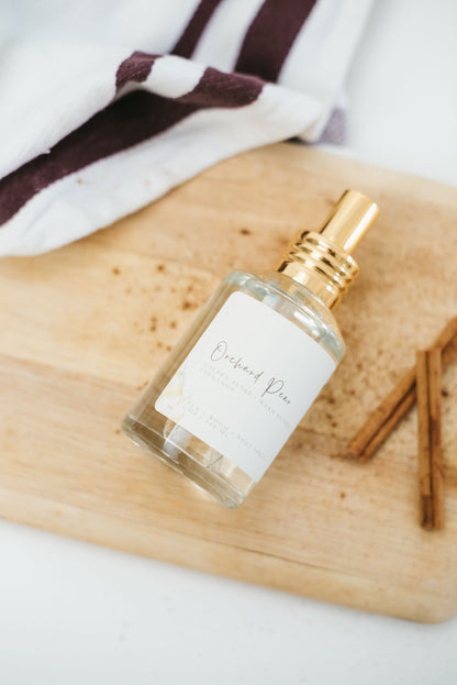 Perfume bottle called Orchard Pear laying on side with a cutting board underneath and cinnamon sticks, ground cinnamon and a towel laying next to the bottle.