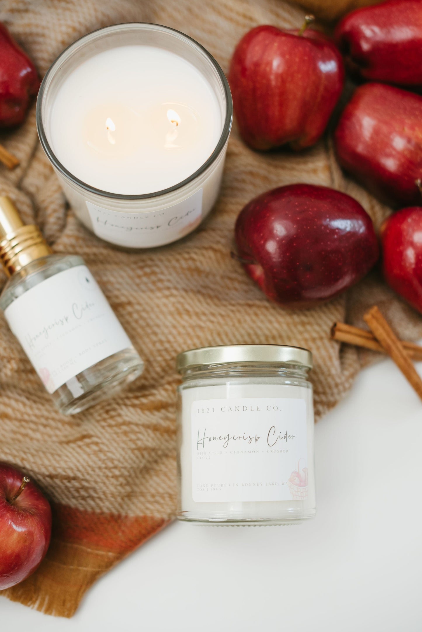 Honeycrisp Cider Candles and linen and room spray laying on top of a woven fall inspired scarf with apples and cinnamon sticks