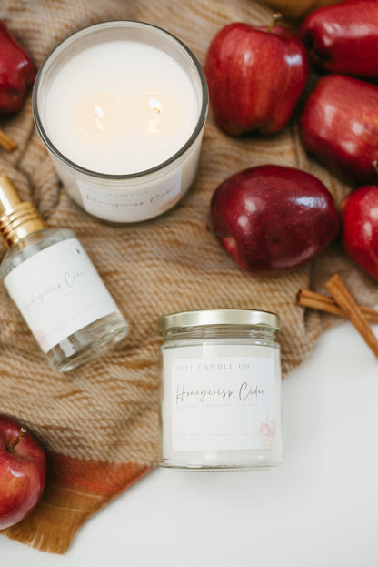 Honeycrisp Cider Candles and linen and room spray laying on top of a woven fall inspired scarf with apples and cinnamon sticks
